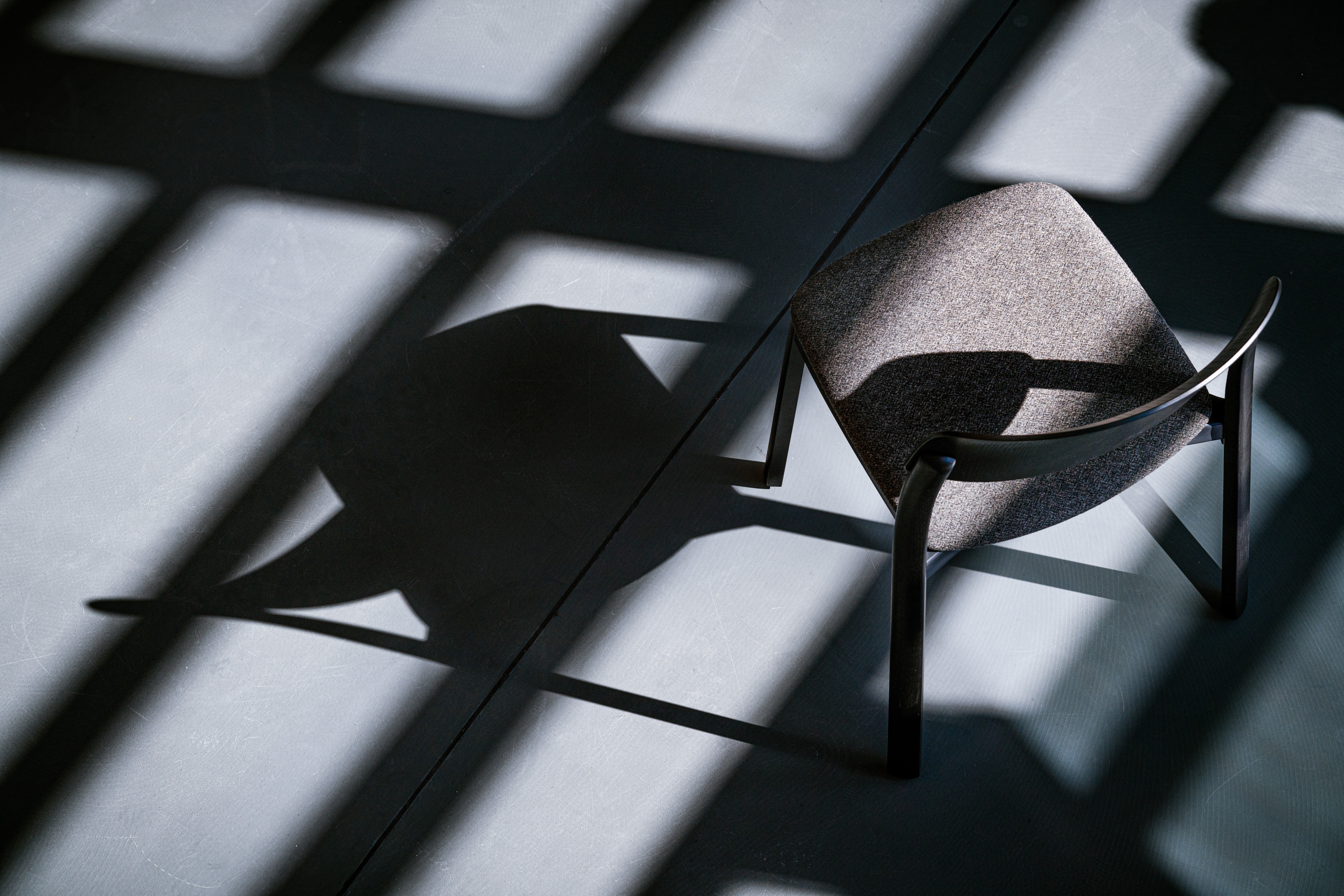 A single  chair with a dark wooden frame and upholstered seat, photographed from above against a light grey floor. Strong directional sunlight casts bold geometric shadows of window frames across the floor, with the chair's own shadow extending dramatically to the left. The high-contrast interplay of light and shadow gives the image a graphic, almost abstract quality.