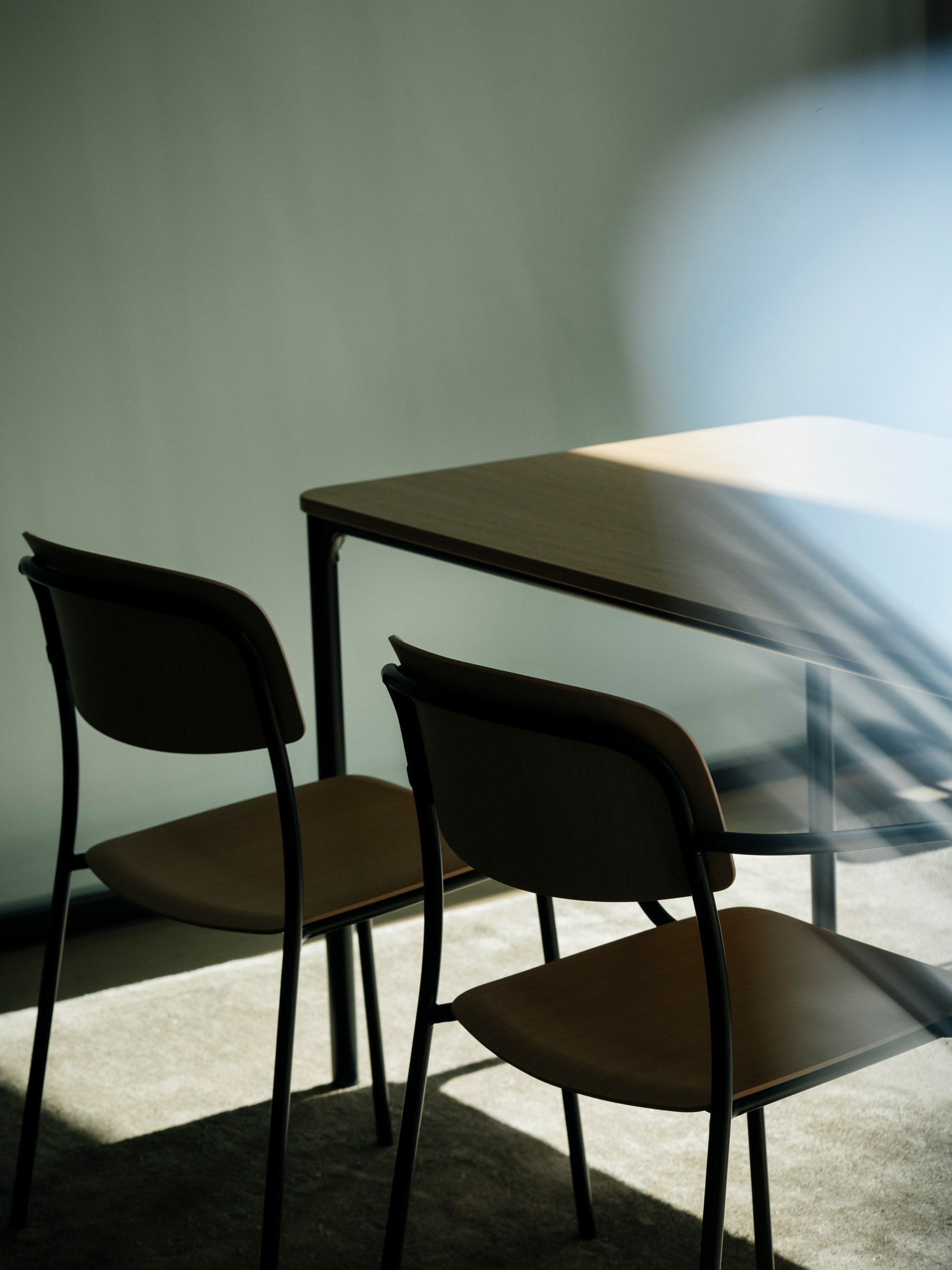 A row of Nate S chairs with dark steel frames and light wood seats and backrests, arranged diagonally on a light wooden floor. Sunlight streams in from the side, casting soft shadows across the floor. A lens flare with rainbow colors adds a dreamy quality to the foreground. More chairs are visible in the background, slightly out of focus.