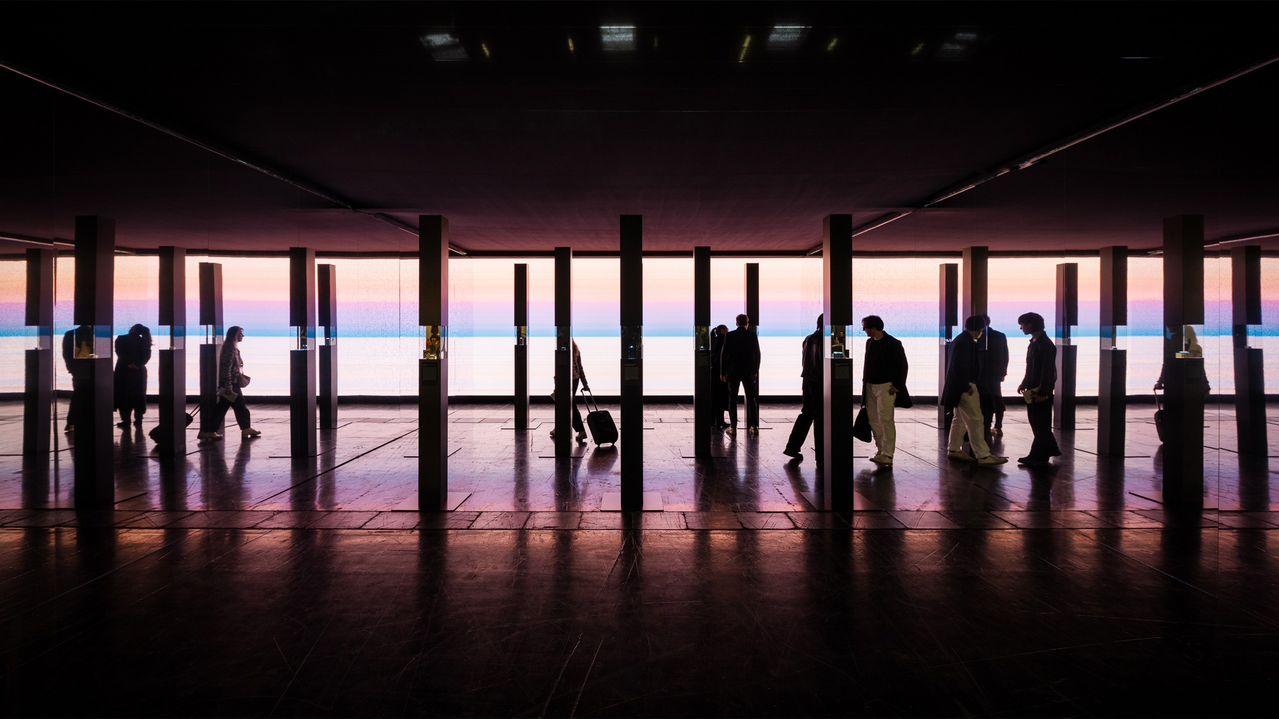 Silhouettes of visitors walking through a darkened exhibition space, framed by a row of tall narrow columns. Behind them, a large-scale video installation displays a panoramic sunset over water in soft shades of pink, blue, and purple. The reflective dark floor mirrors the atmospheric light, creating a dramatic and immersive scene.