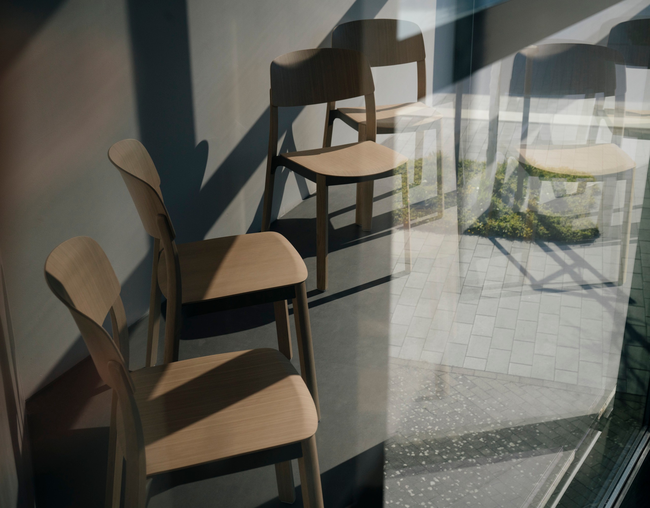 Five wooden chairs are freely positioned in a space behind a glass window. They are photographed through the glass, so that reflections are visible in the image overlaying the chairs.