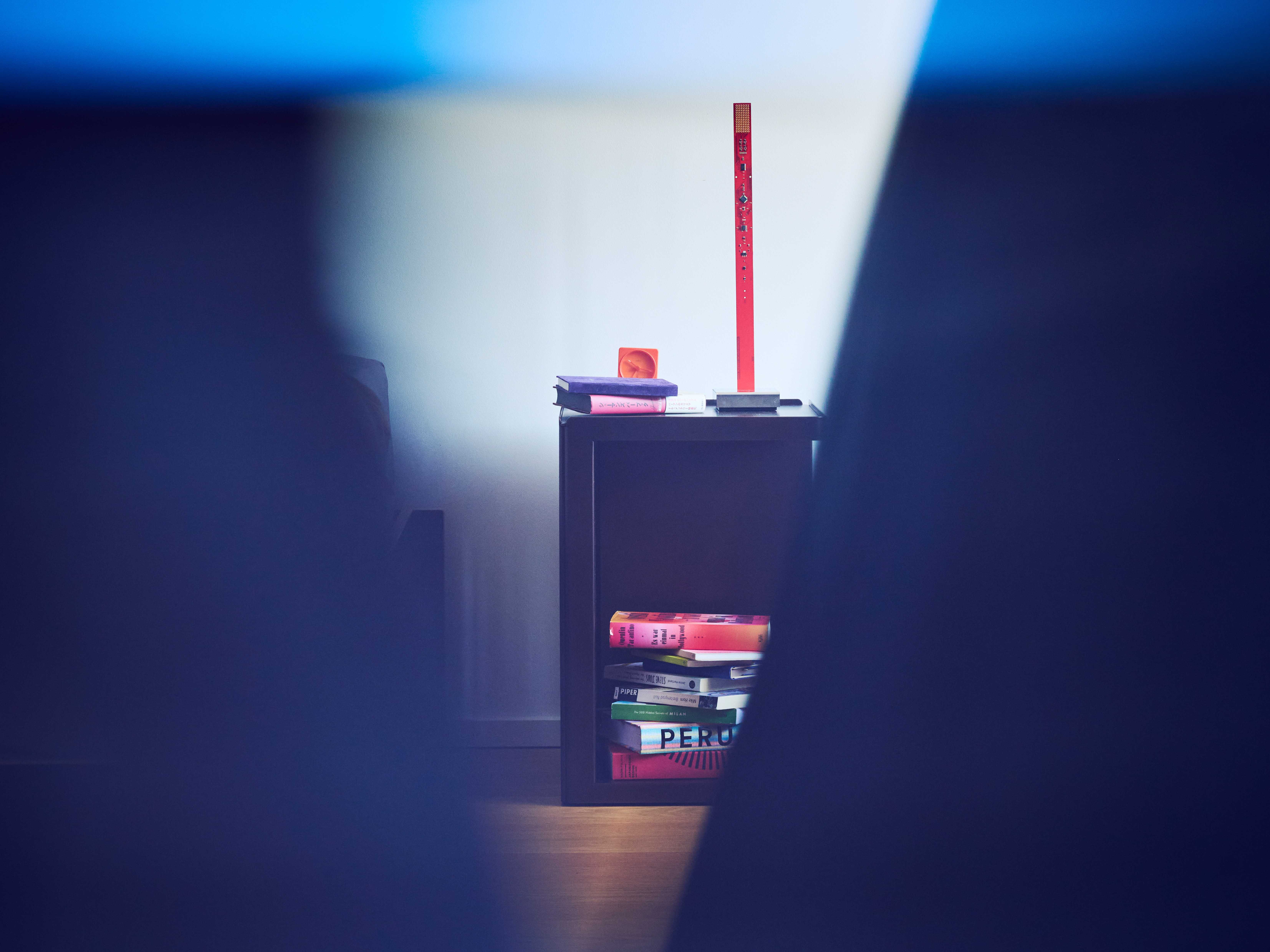 A moody interior shot framed by out-of-focus dark shapes in the foreground, revealing a small dark side table in the background. The table holds a stack of colorful books — including one titled 'Peru' — topped with a tall slim red lamp. The scene is bathed in cool blue ambient light, with a narrow beam of white light illuminating the side table at the center.