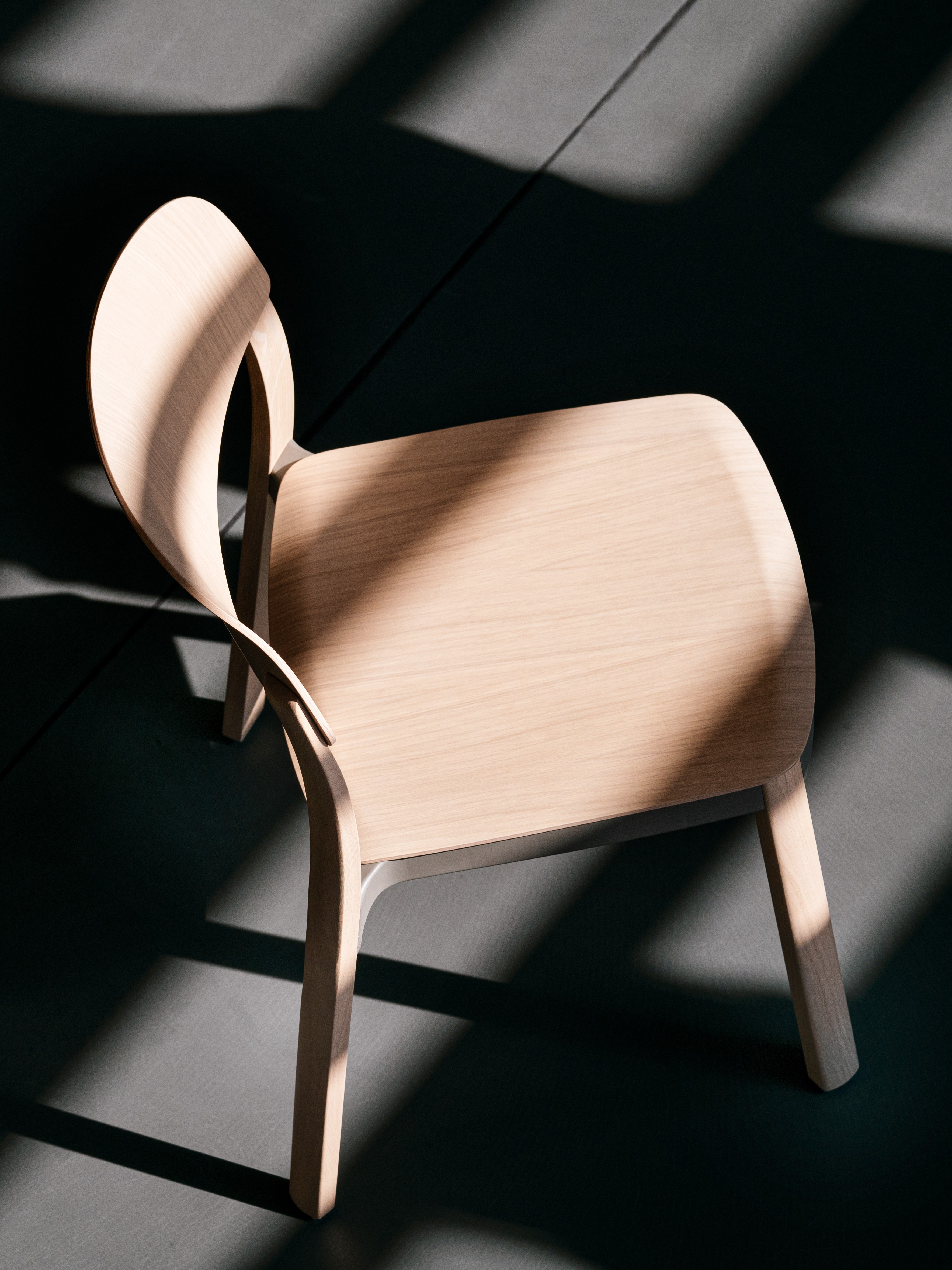 A natural wooden chair photographed from above, standing on a concrete floor. The sun ist casting hard shadows of the architecture and the chair on the ground.