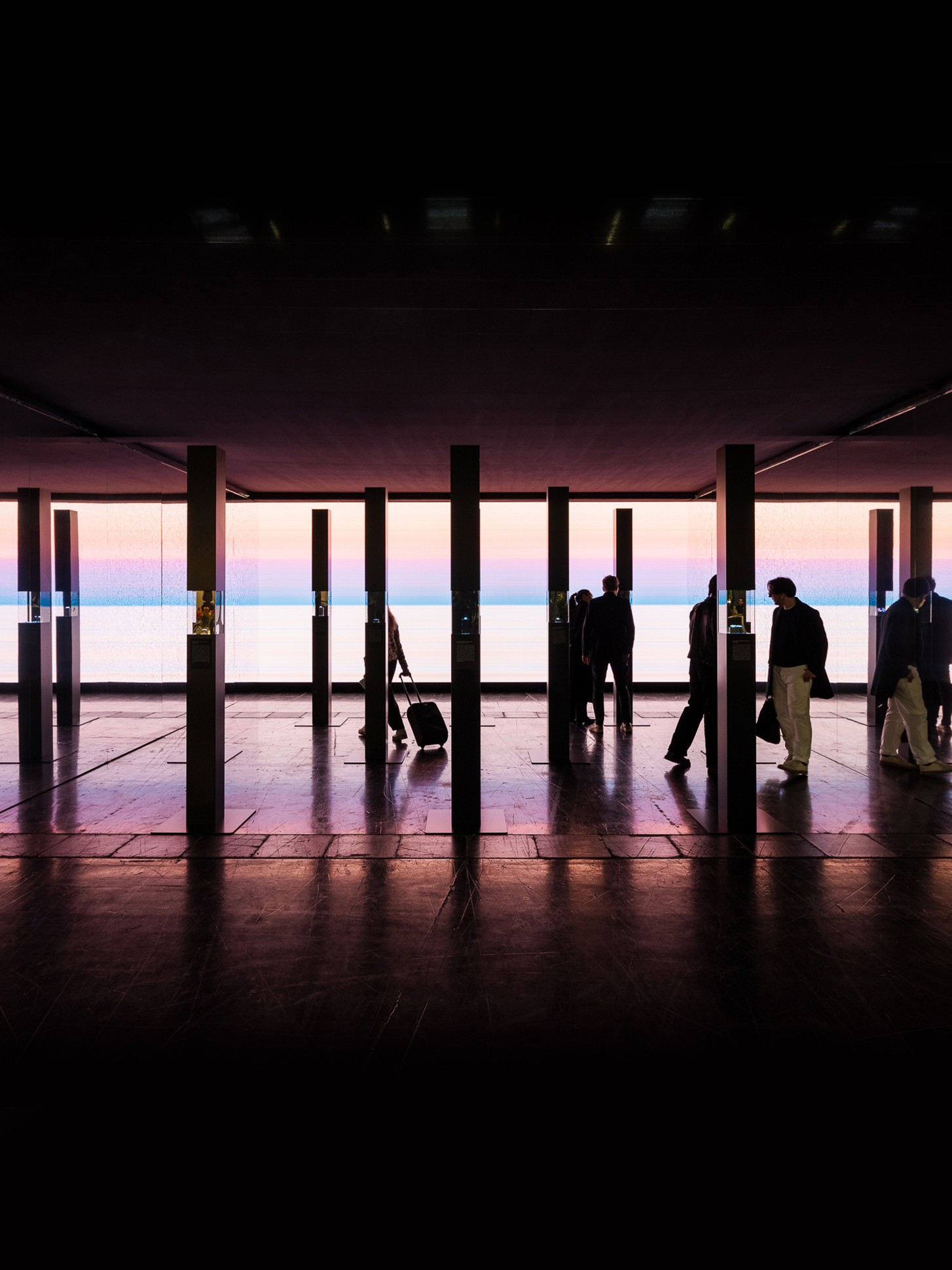 Silhouettes of visitors walking through a darkened exhibition space, framed by a row of tall narrow columns. Behind them, a large-scale video installation displays a panoramic sunset over water in soft shades of pink, blue, and purple. The reflective dark floor mirrors the atmospheric light, creating a dramatic and immersive scene.