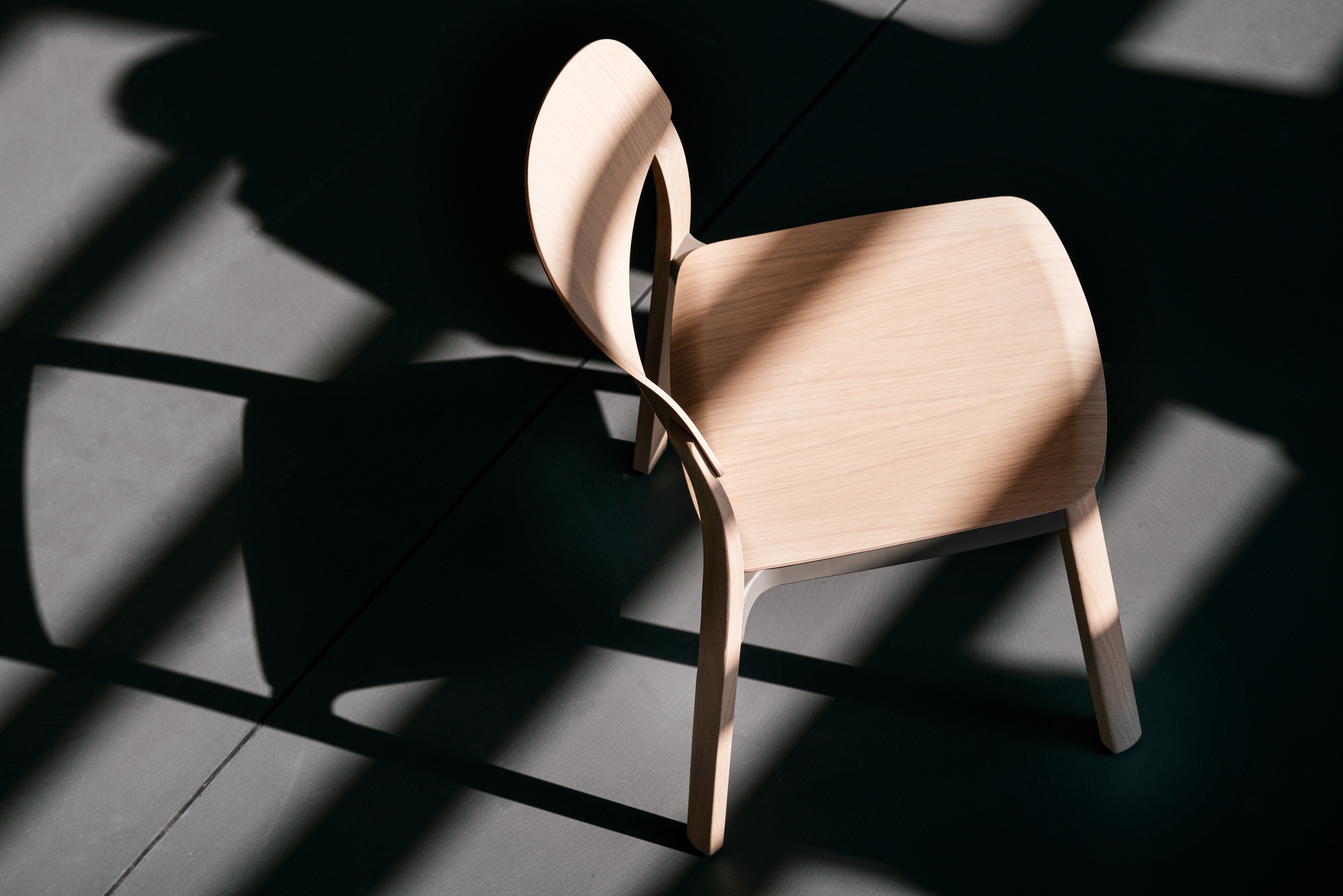 A natural wooden chair photographed from above, standing on a concrete floor. The sun ist casting hard shadows of the architecture and the chair on the ground.