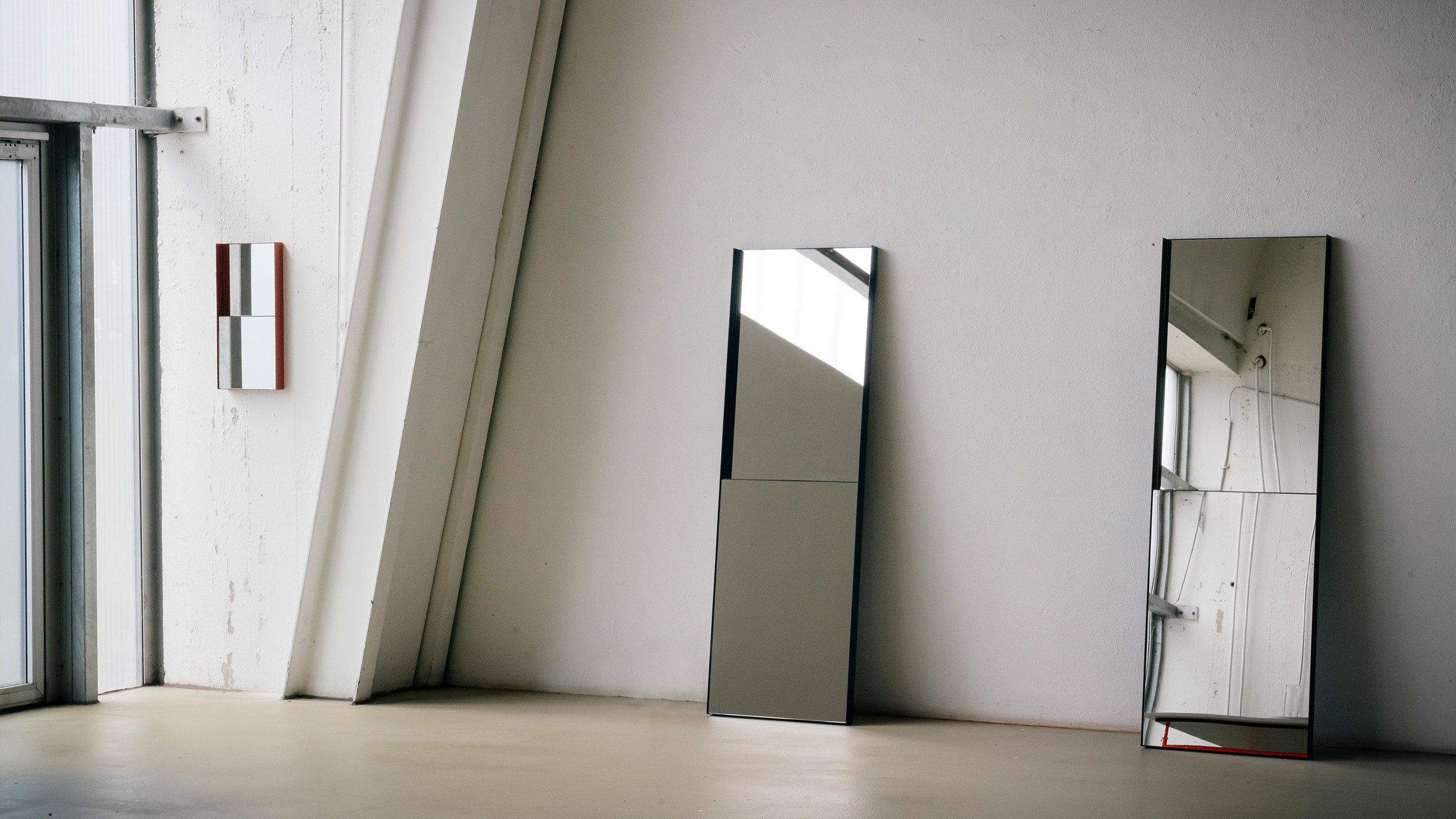 Three mirrors displayed in a bright, raw industrial space with white walls and a concrete floor. Two tall floor mirrors with thin dark frames lean against the wall, each divided into two separate mirror panels within one frame. A smaller wall-mounted mirror with a terracotta-colored frame hangs on the left, featuring the same two-panel design in a compact format. All three mirrors reflect the surrounding space and incoming light from the large windows on the left.