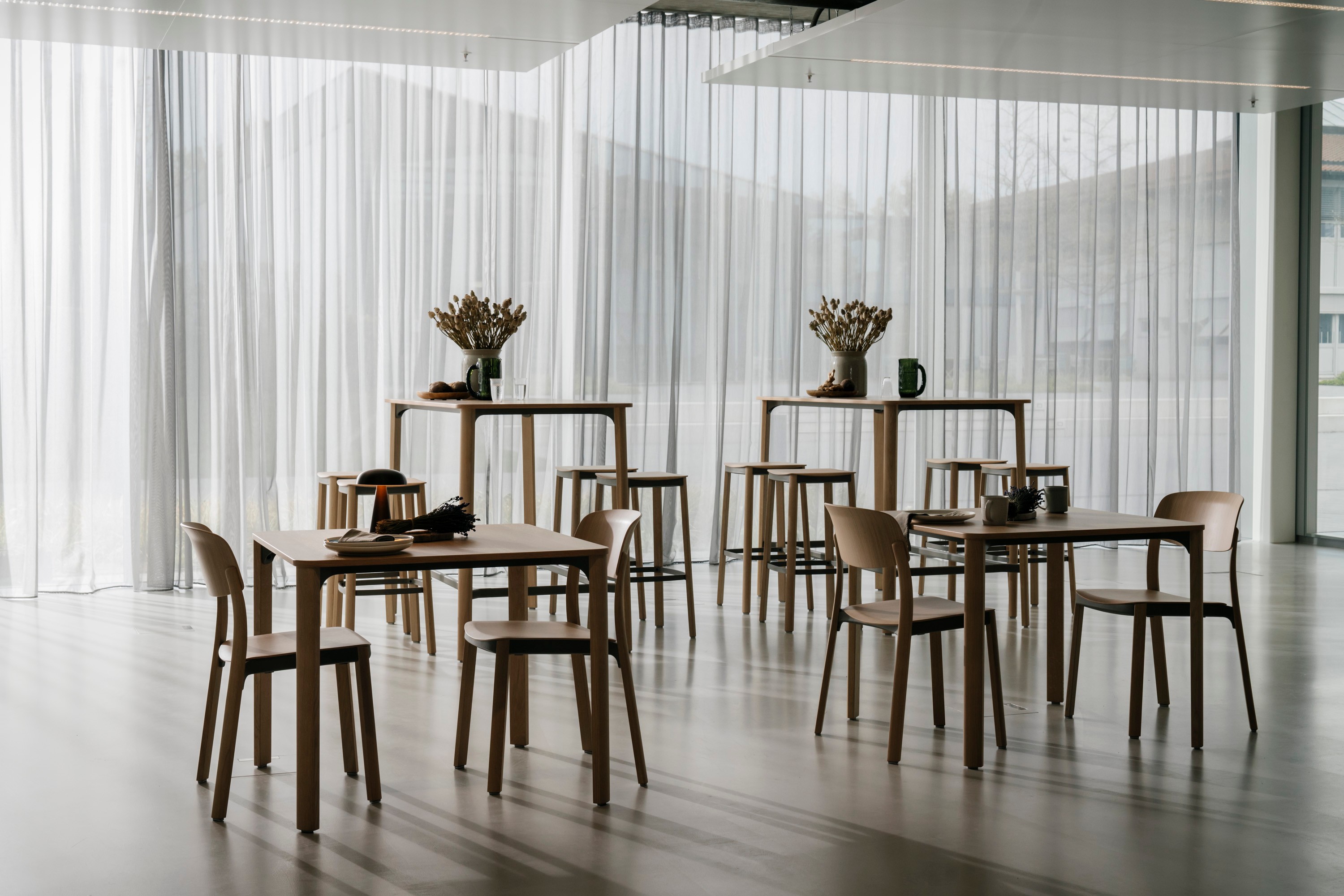 A bright, minimalist interior setting featuring multiple natural oak dining tables and matching wooden chairs arranged across a light wooden floor. Taller bistro-style tables with stools are placed in the background. The tables are styled with dried flower arrangements, ceramic vessels, and green glassware. Floor-to-ceiling sheer white curtains diffuse the daylight and reveal a landscape with hills beyond. The overall atmosphere is calm and Scandinavian-inspired.