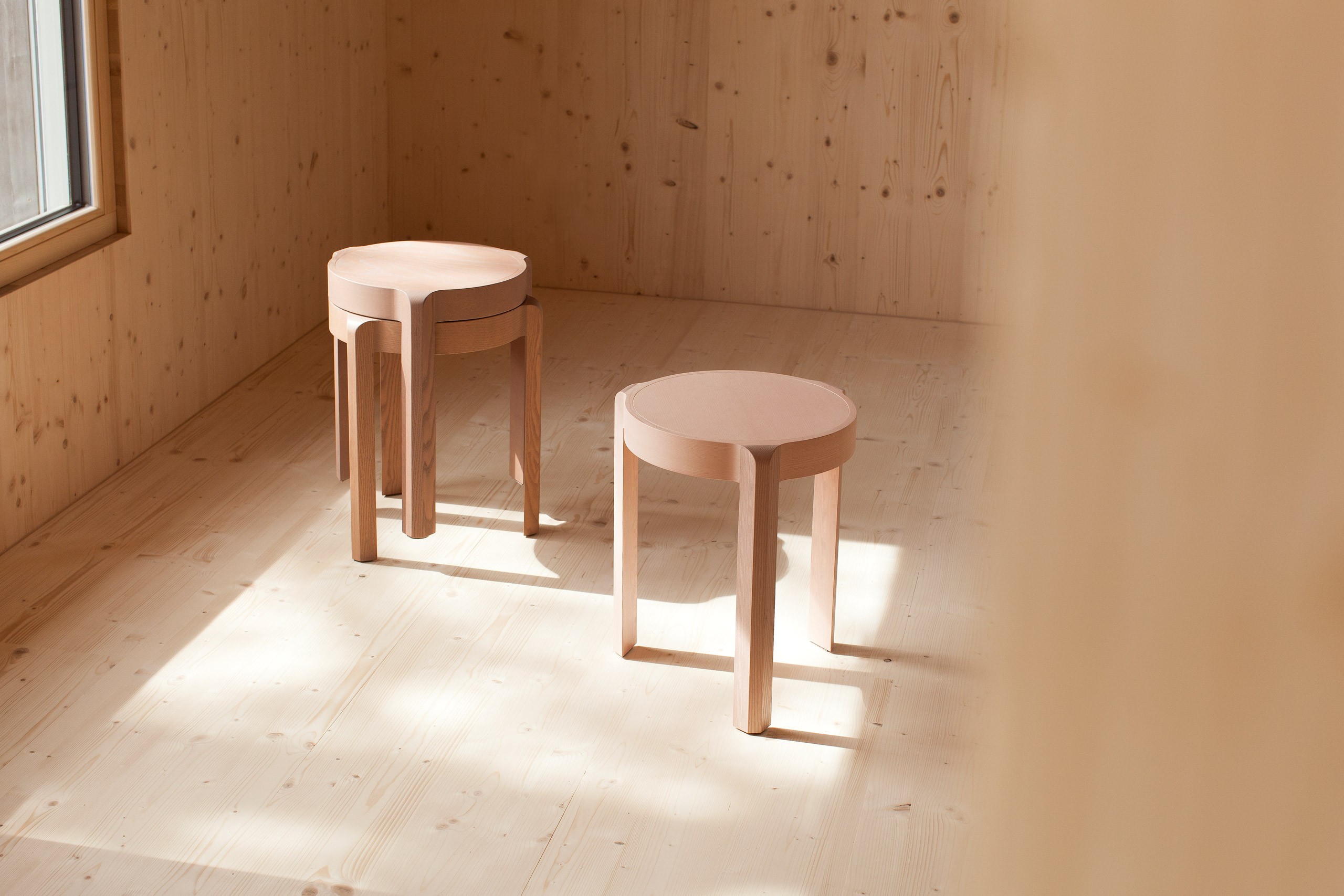 Three light wood stackable stools in a bright corner room with natural pine wood walls and floor. One stool stands alone in the foreground, while two identical stools are stacked on top of each other in the background. The stools feature round tops and three curved legs. Soft sunlight enters through a window to the left, casting gentle shadows across the floor and highlighting the natural grain of both the furniture and the surrounding interior.