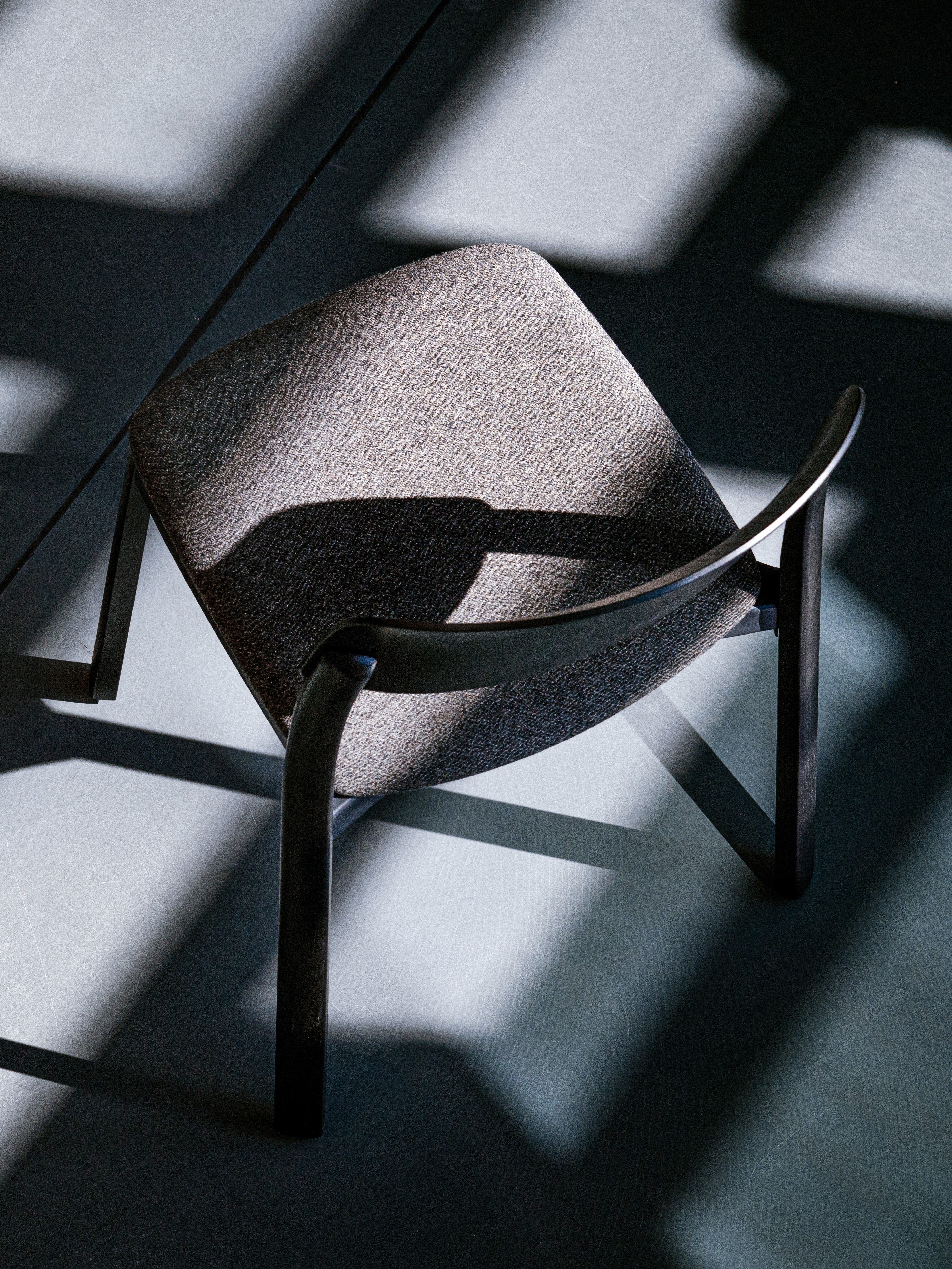 A single chair with a dark wooden frame and upholstered seat, photographed from above against a light grey floor. Strong directional sunlight casts bold geometric shadows of window frames across the floor, with the chair's own shadow extending dramatically to the left. The high-contrast interplay of light and shadow gives the image a graphic, almost abstract quality.