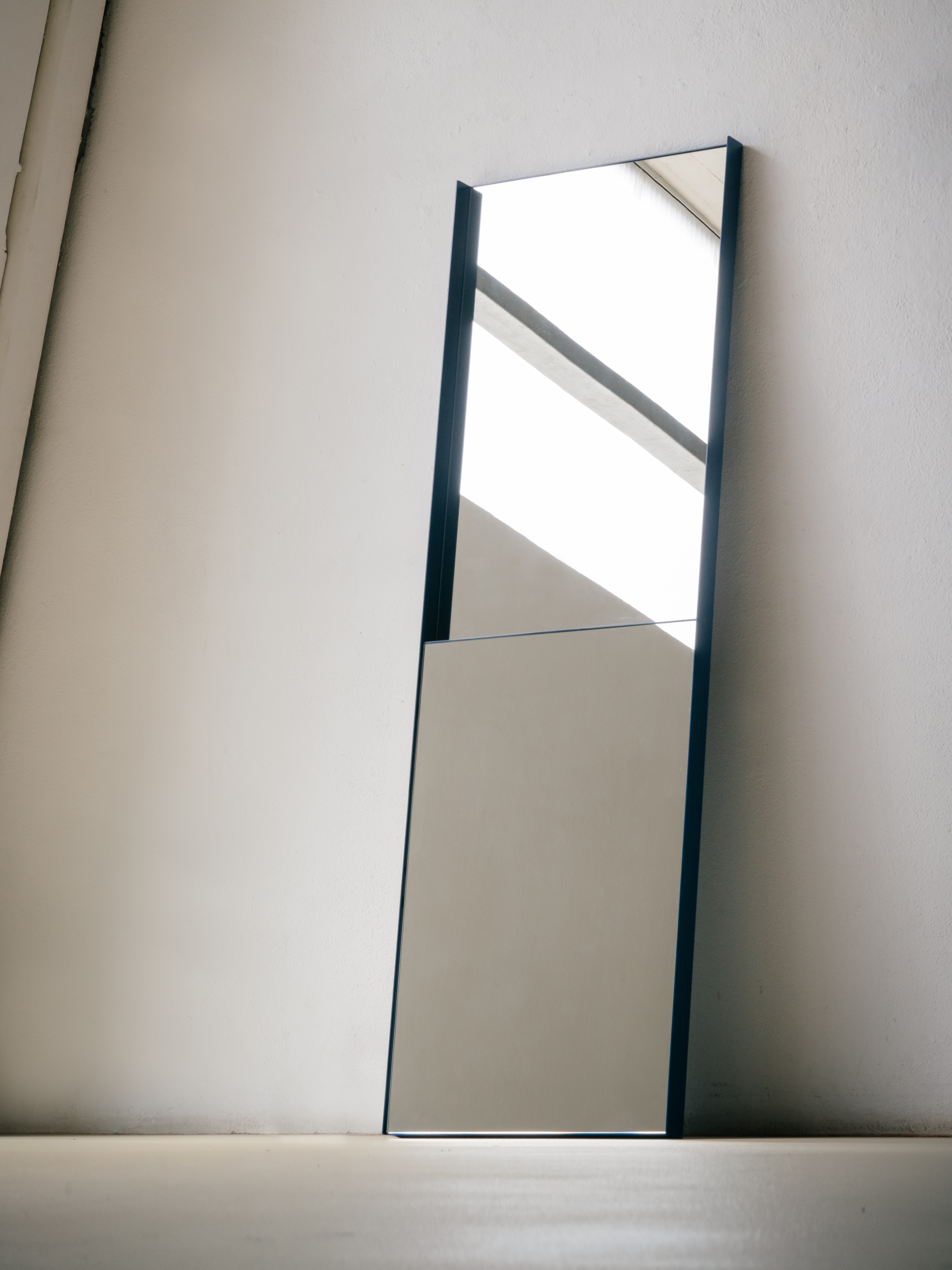 Three mirrors displayed in a bright, raw industrial space with white walls and a concrete floor. Two tall floor mirrors with thin dark frames lean against the wall, each divided into two separate mirror panels within one frame. A smaller wall-mounted mirror with a terracotta-colored frame hangs on the left, featuring the same two-panel design in a compact format. All three mirrors reflect the surrounding space and incoming light from the large windows on the left.
