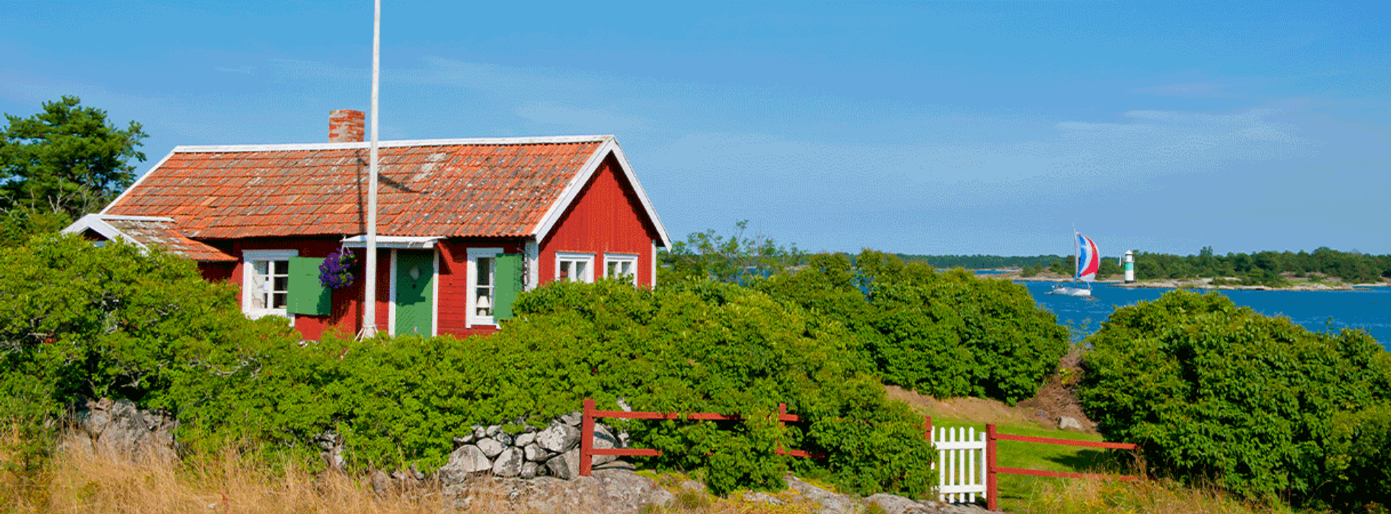 En röd sommarstuga med vita knutar och gröna dörrar som ligger vid havet.