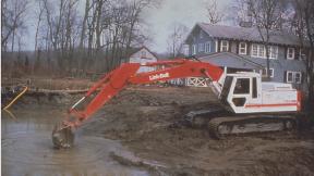 A Link-Belt excavator working in a small pond near a blue house