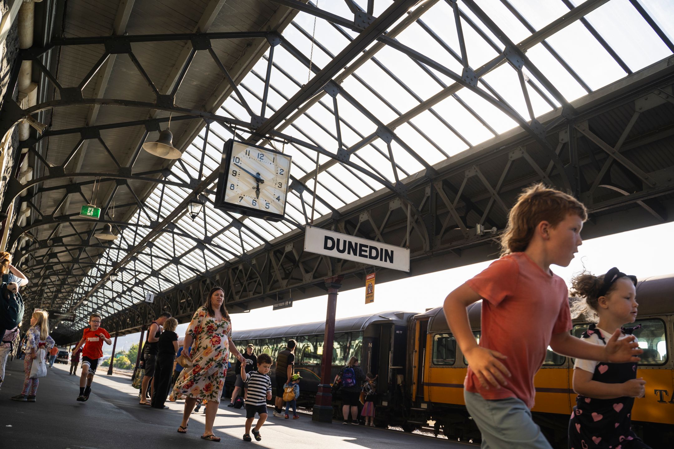 A bustling scene at Dunedin train station featuring families and children running along the platform. A large clock hangs above, showing the time. The station has a vintage design with a mix of passengers, highlighting the lively atmosphere of travel in Dunedin.