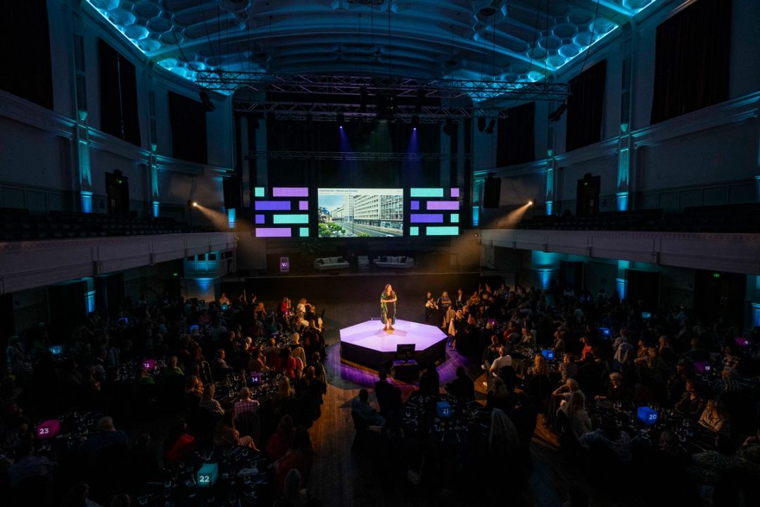 Person in centre of the town hall on a plinth speaking to a room full of attentive people at tables.