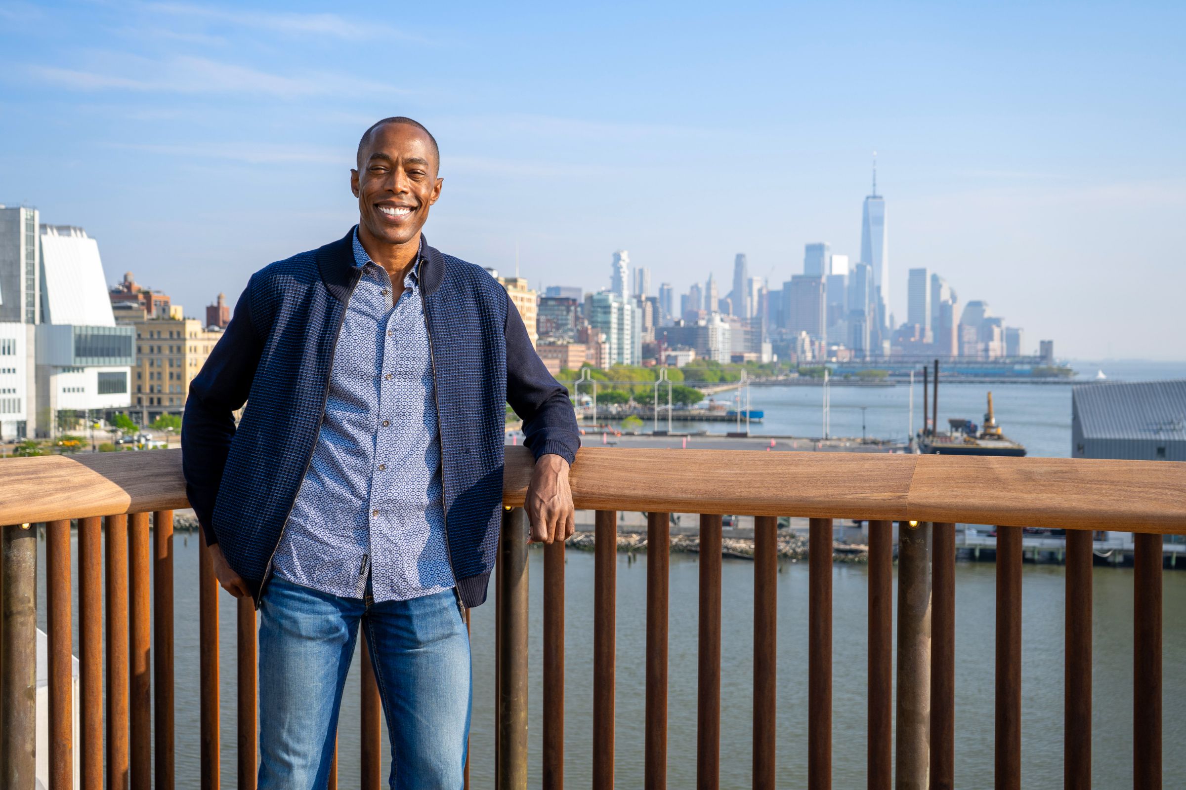 A man leaning against a wooden railing with the water and city skyline in the background