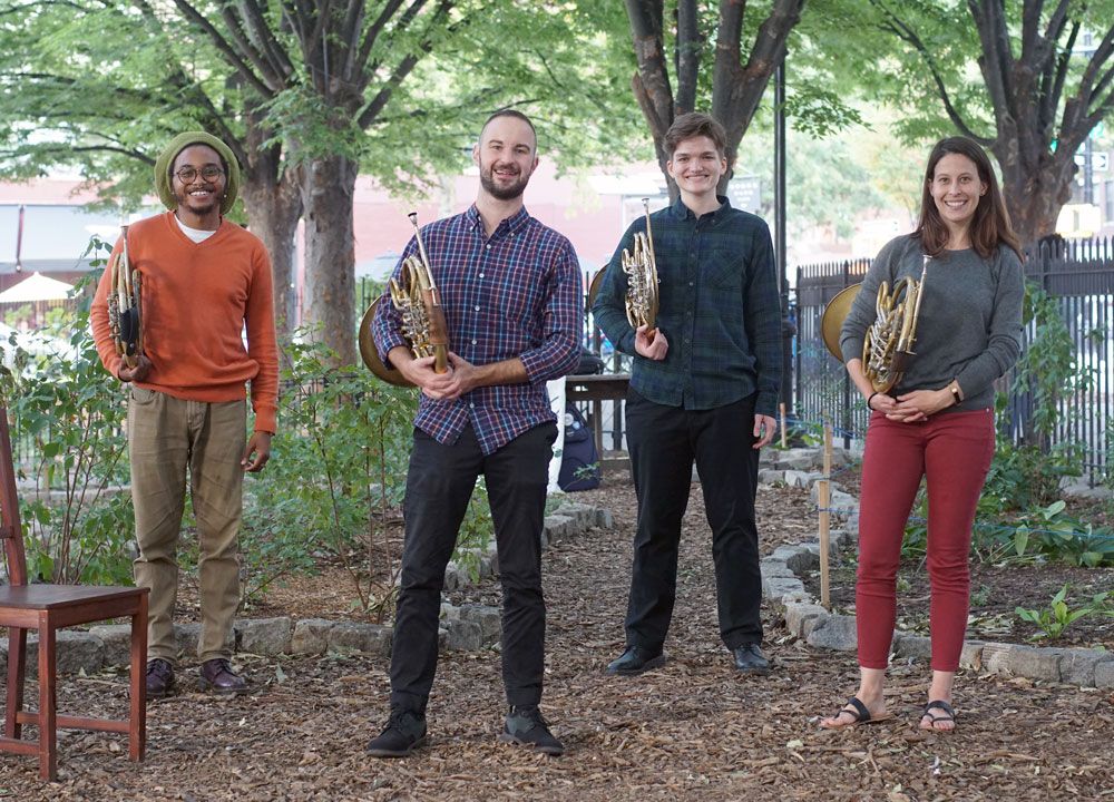 Four people hold french horns in a park.