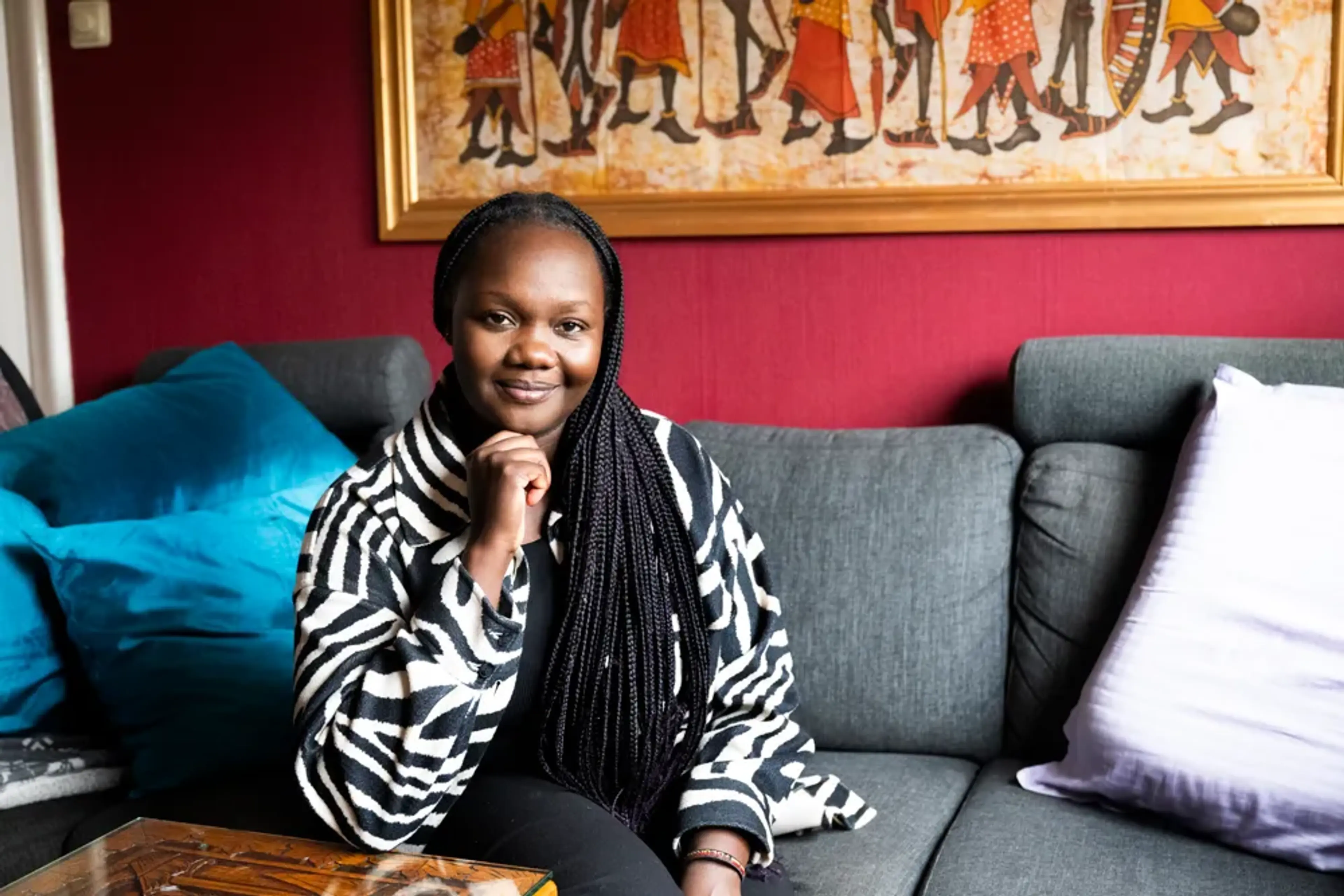 Faith sitter i en soffa med en hand vilande mot hakan. Hon har en zebramönstrad jacka. I bakgrunden syns en färgstark tavla med afrikanska motiv och en vinröd vägg.
