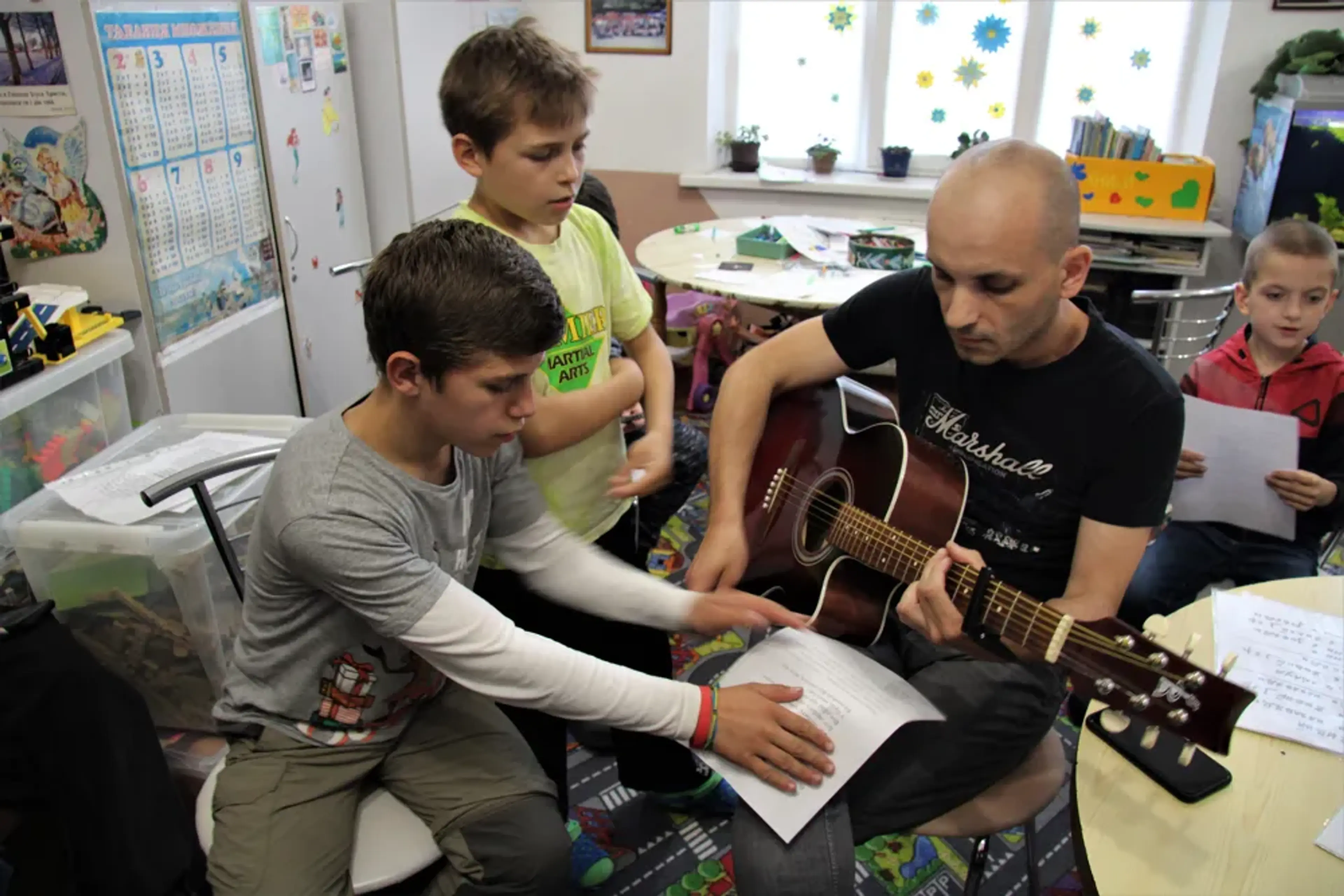 En man visar hur man spelar gitarr för två unga pojkar. De hjälper honom med noterna.