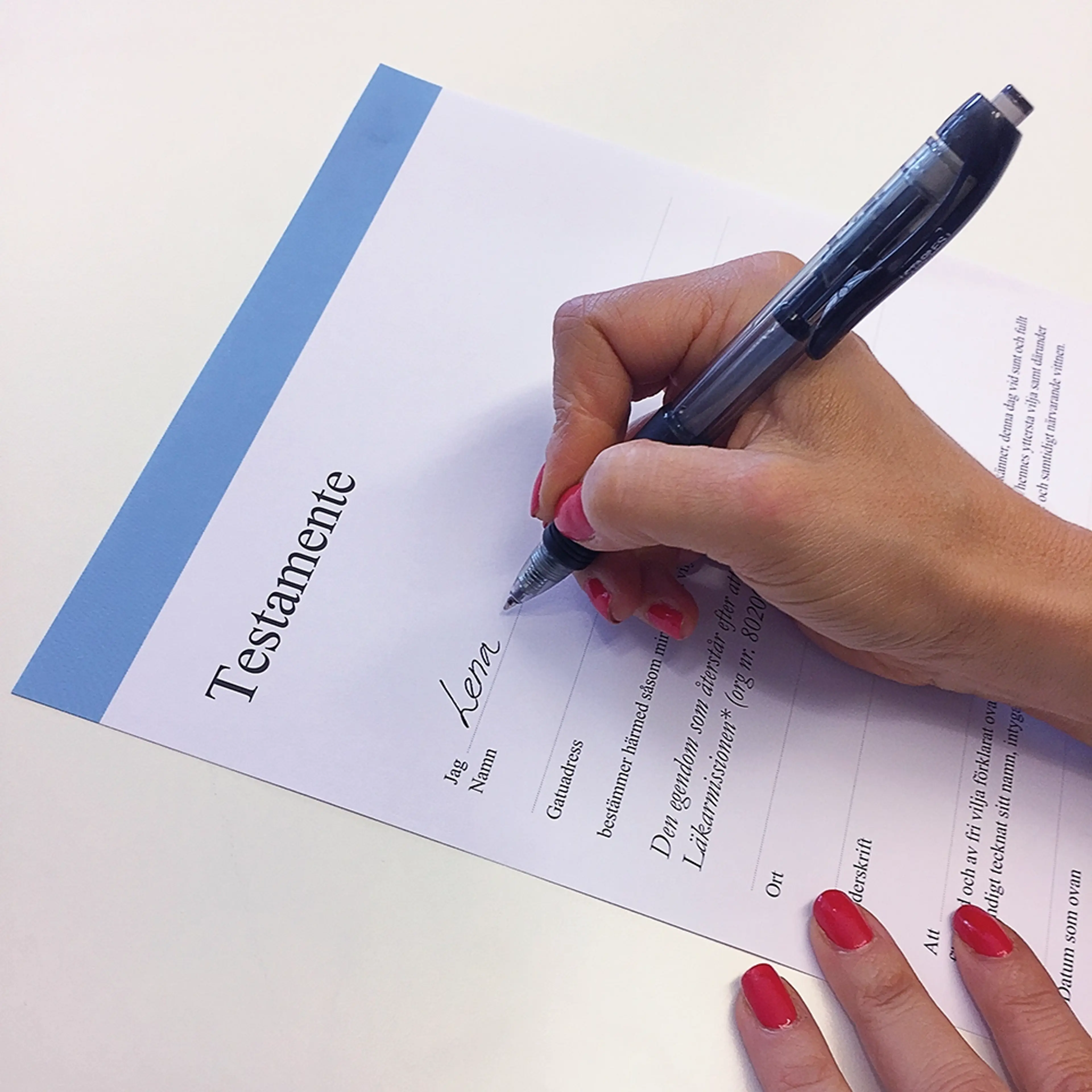 En person med rödmålade naglar skriver under ett testamente med en svart penna. Dokumentet har en blå kant upptill och rubriken ”Testamente” syns tydligt.