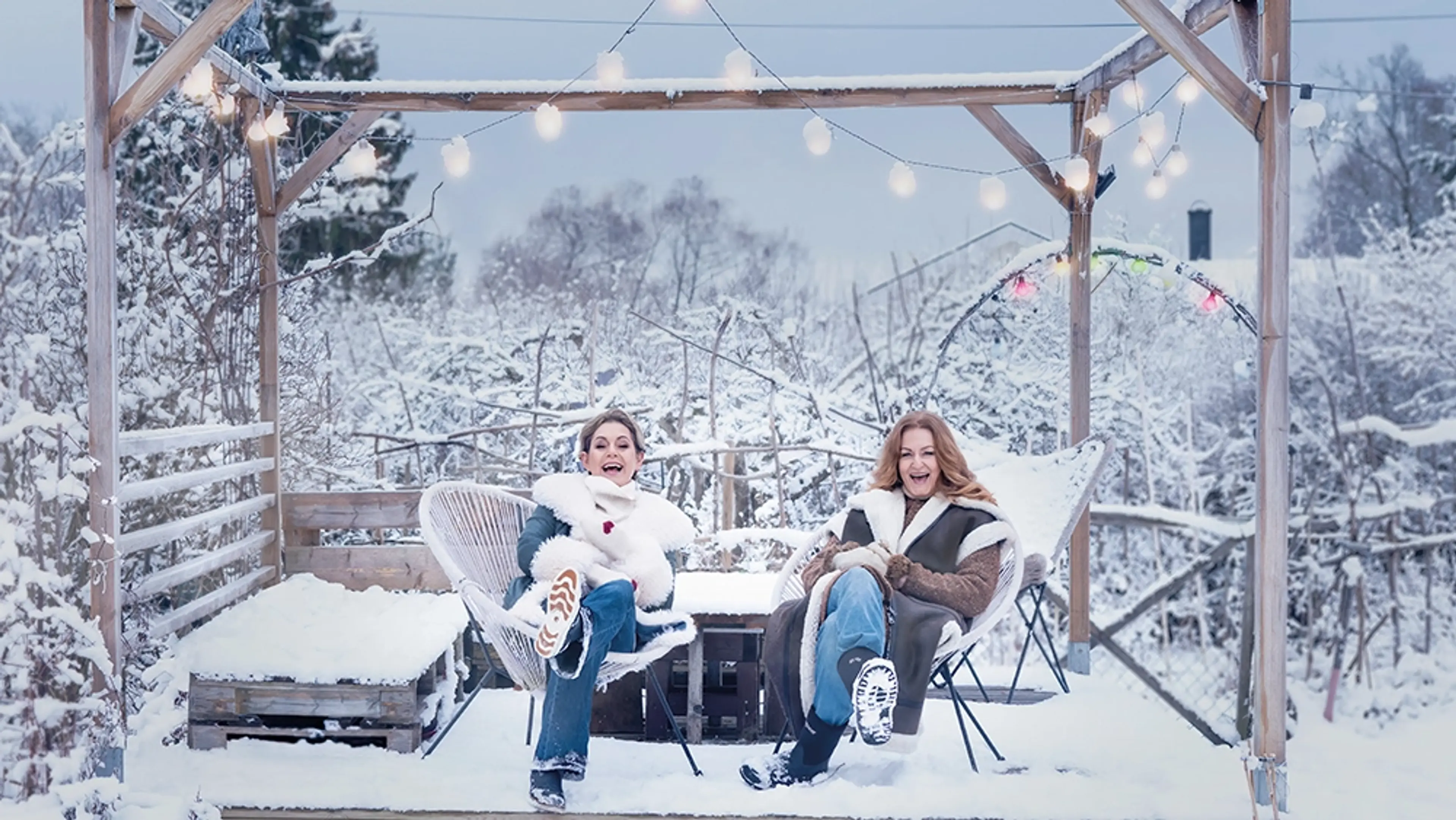 Helen Sjöholm och Anna Stadling sitter utomhus i en kall omgivning fylld med snö. Dom sitter i varsin stol och skrattar.