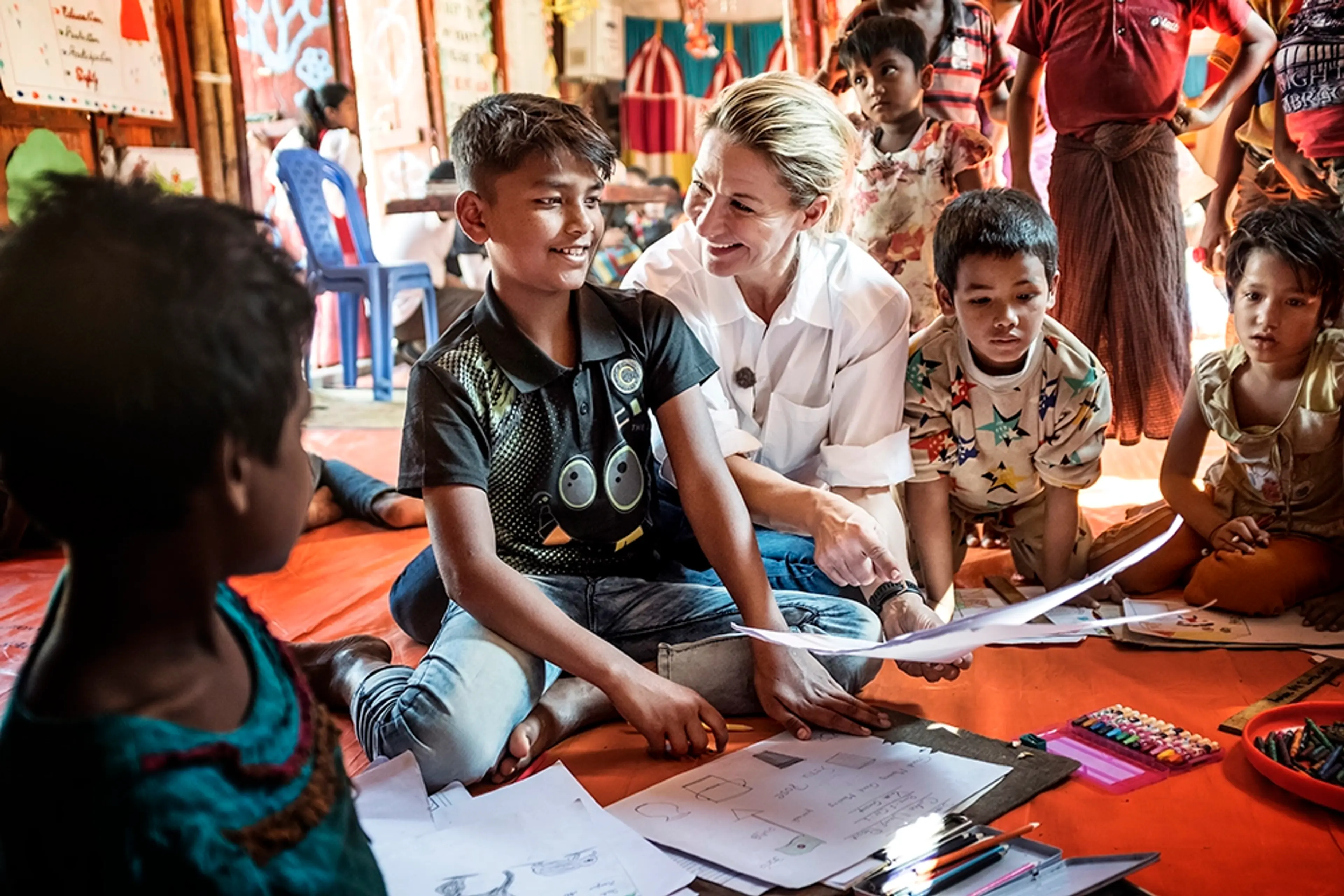 Kristin sitter på en orange matta tillsammans med några barn på ett flyktingläger i Bangladesh