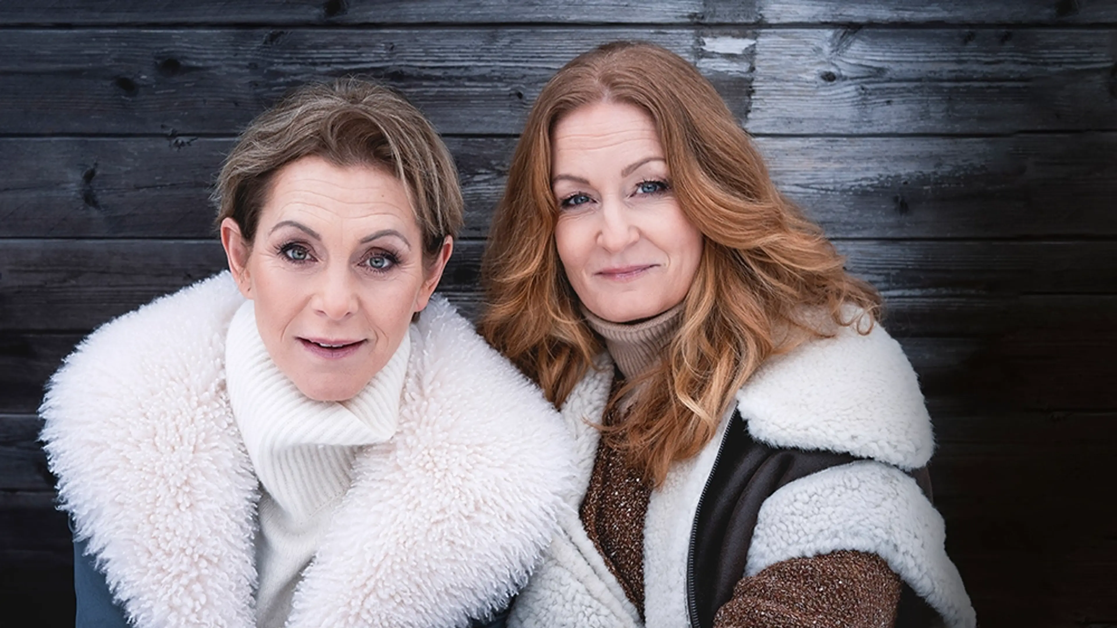 Helen Sjöholm och Anna Stadling står bredvid varandra framför en grå trävägg. Dem har på sig vinterkläder.