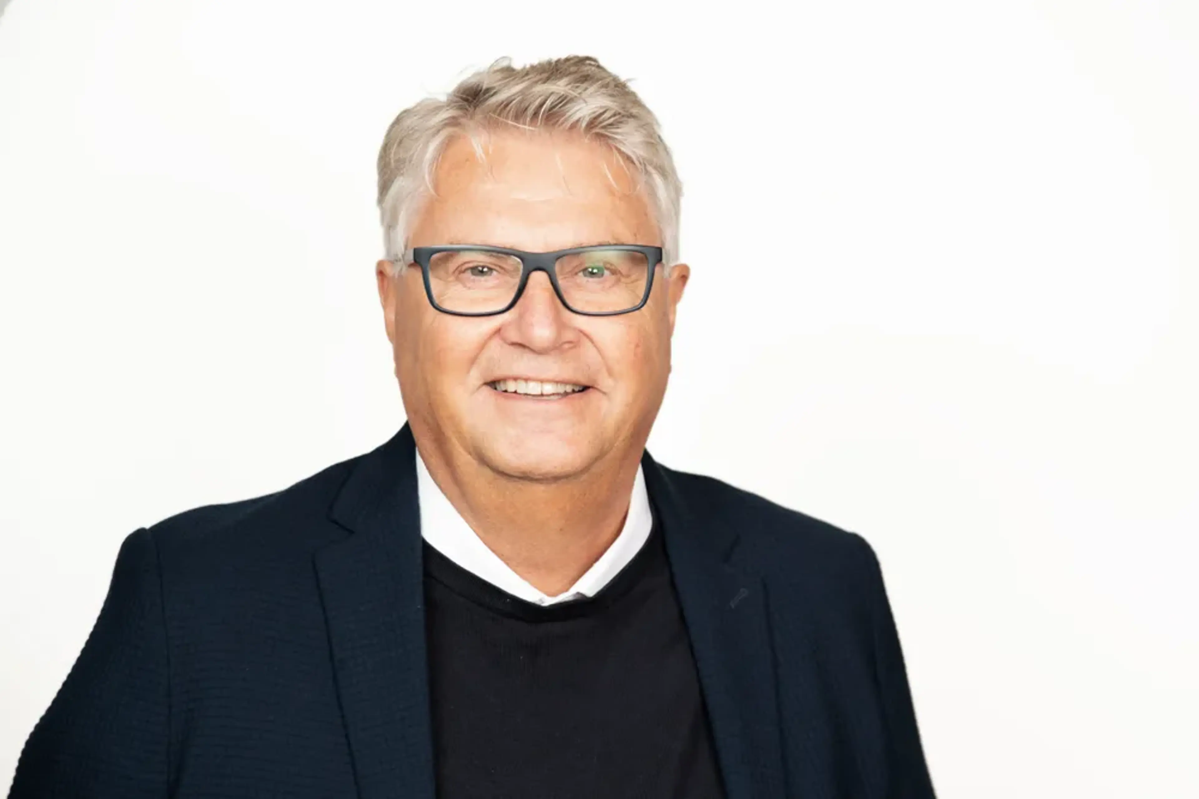 Hans Hallström står framför en vit bakgrund. Han har på sig kostym och ler in i kameran.