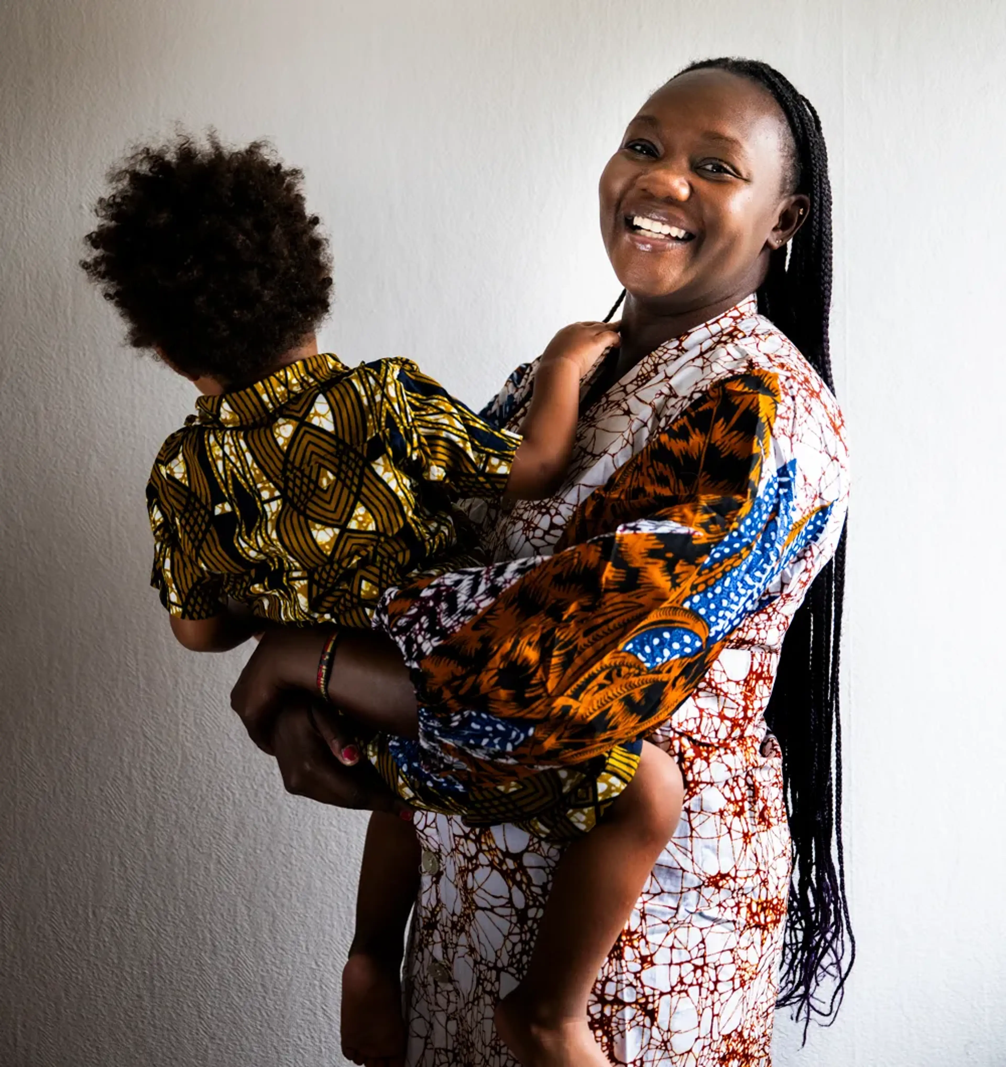 Faith Kakembo håller sitt barn i famnen. Båda bär färgstarka kläder med traditionella afrikanska mönster. De står mot en ljus vägg, och kvinnan ser rakt mot kameran med ett varmt uttryck.