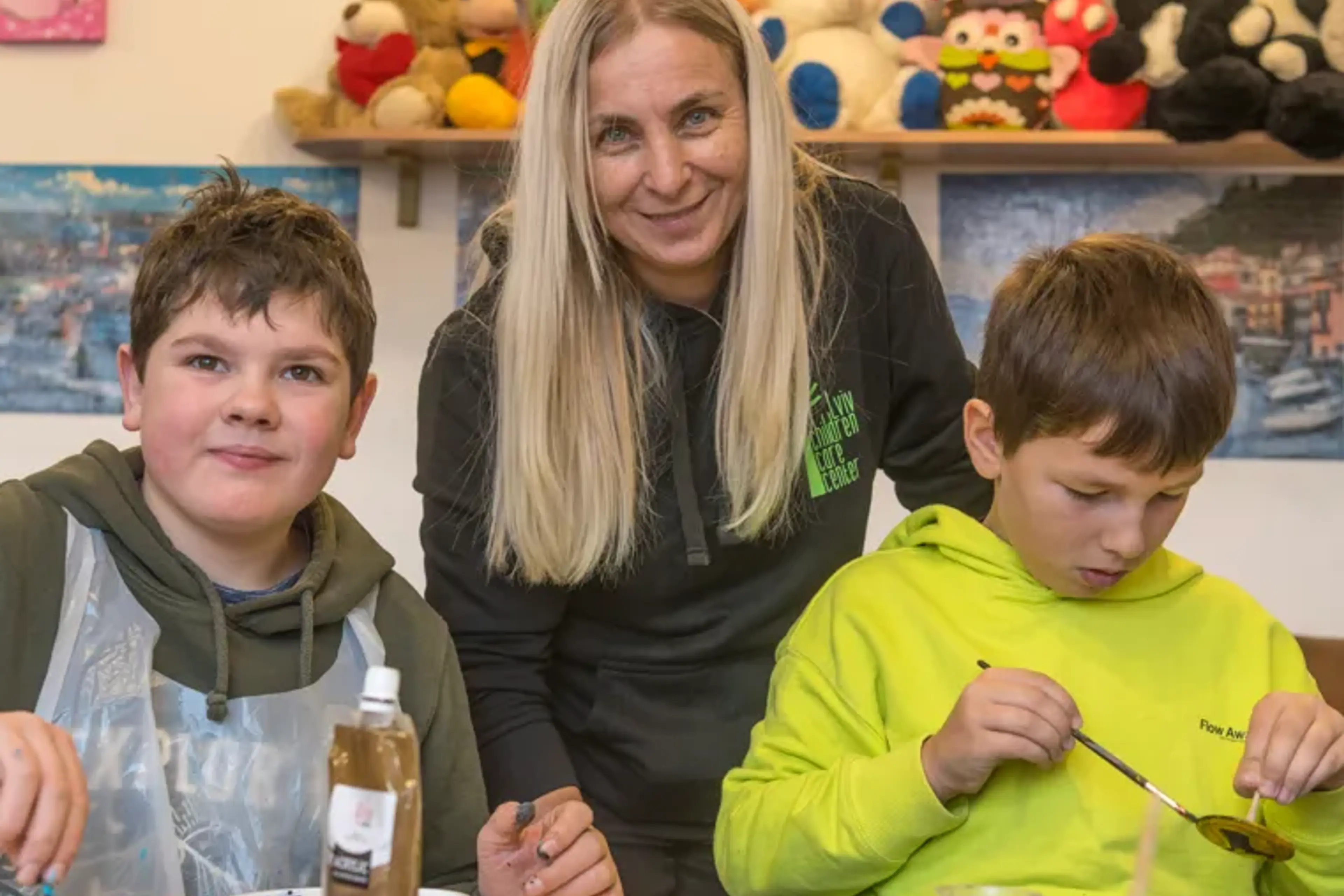 Vera står mellan två unga pojkar som sitter och målar