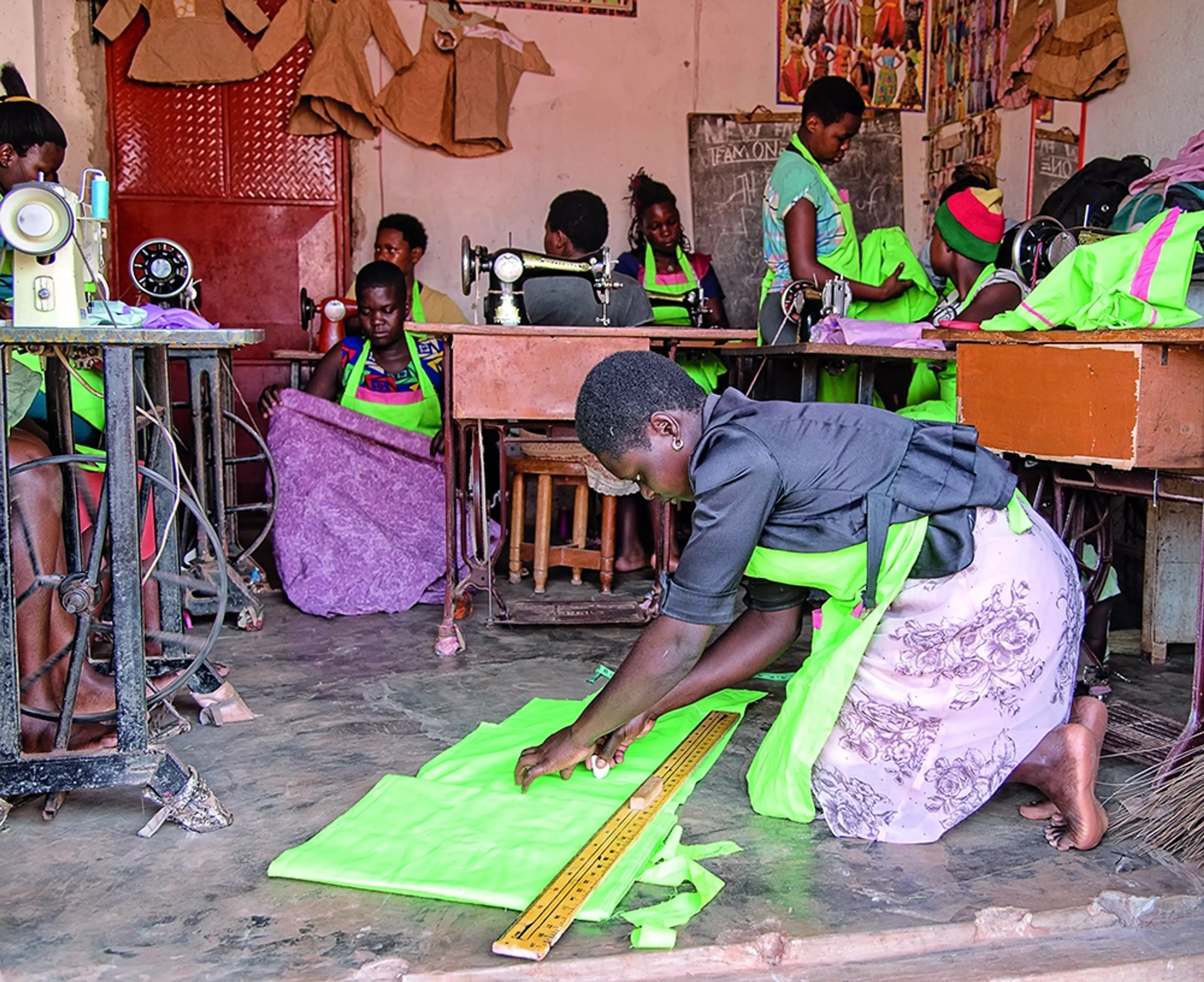 Flera kvinnor syr kläder tillsammans i en mörk lokal. Alla kvinnor har på sig limegröna förkläden.