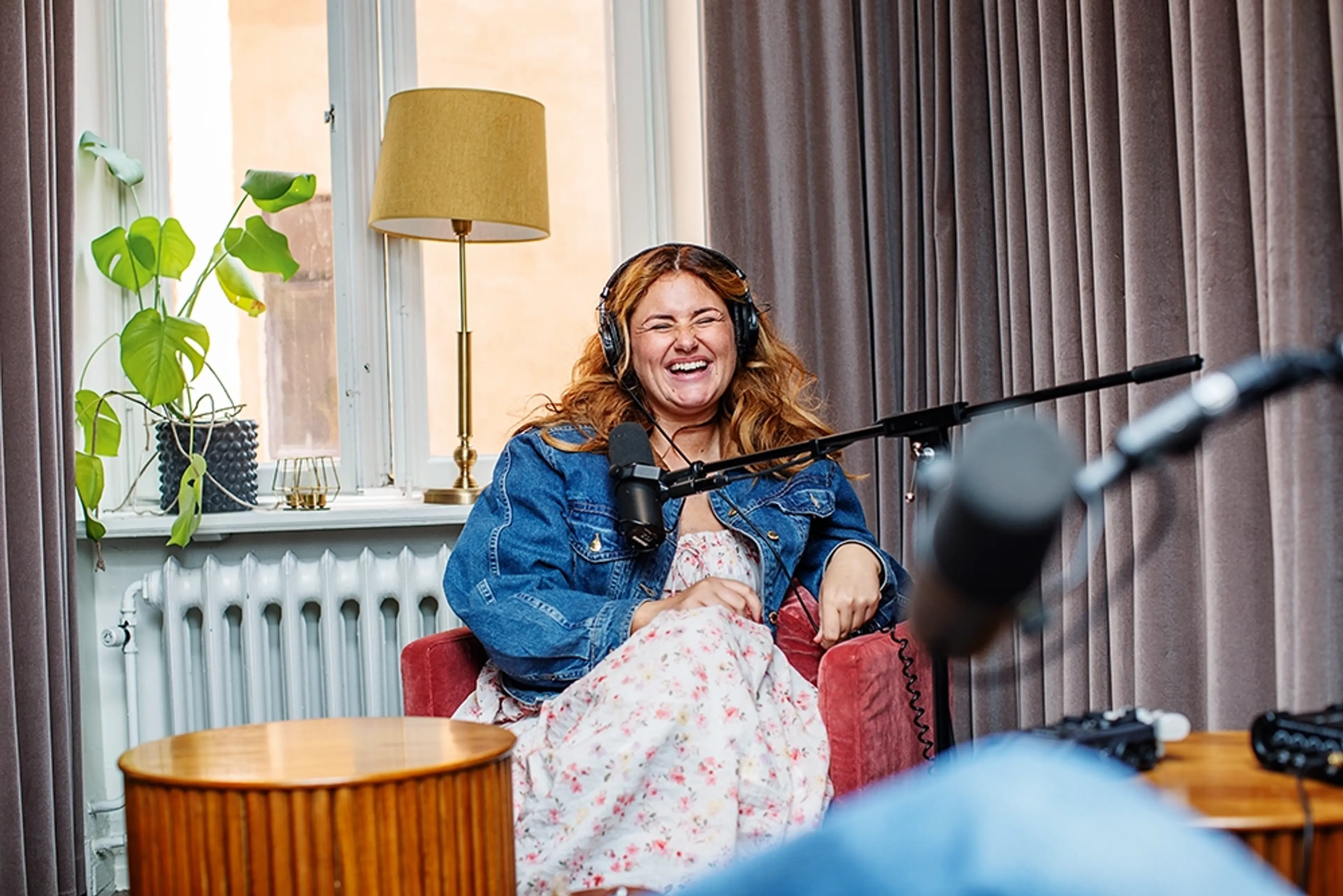 En kvinna sitter i en fåtölj framför en mikrofon, skrattar och har hörlurar på sig. Hon bär en blommig klänning och en jeansjacka. Rummet har stora fönster med gardiner, en grön växt i fönstret och en bordslampa i bakgrunden.