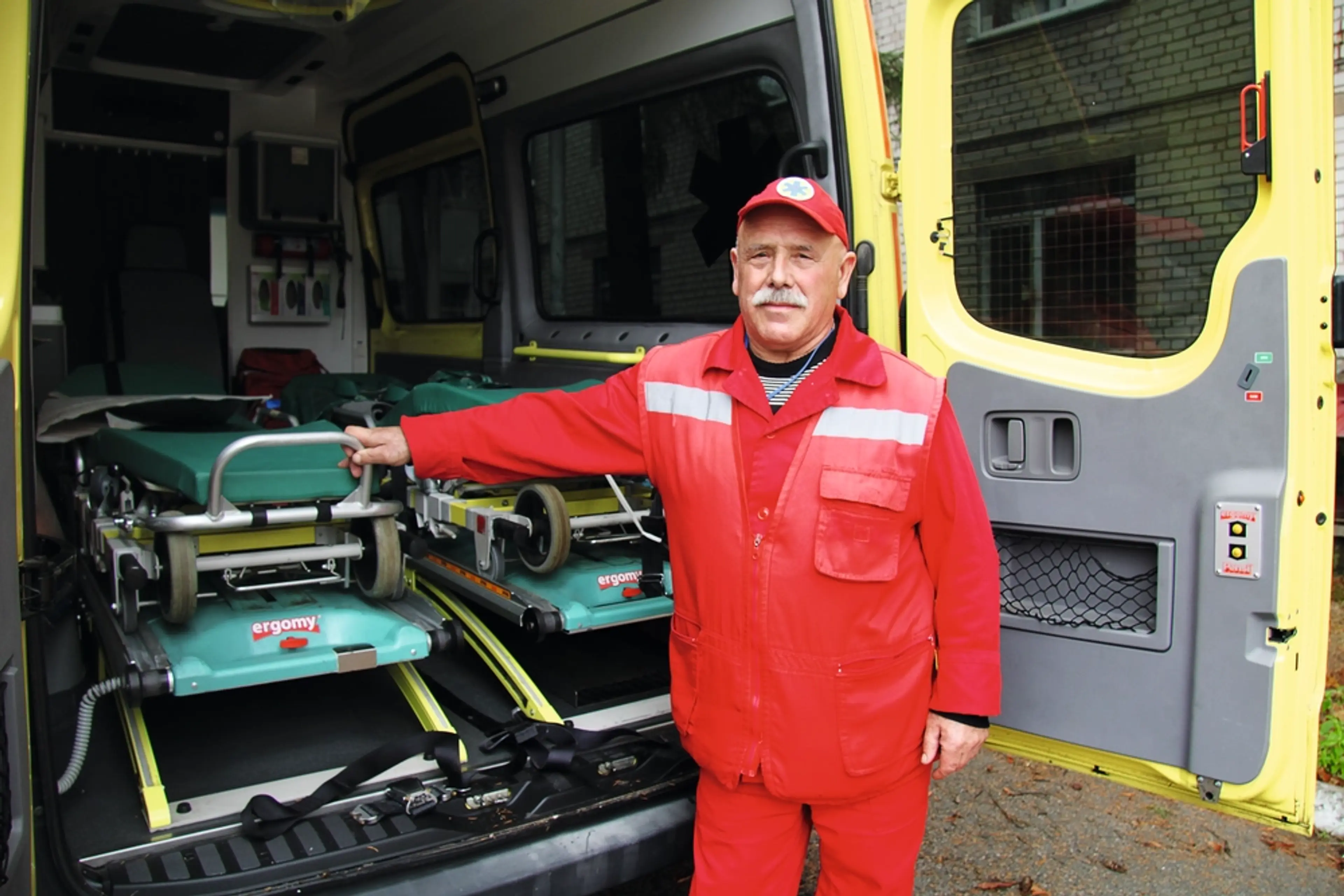Ambulanssjukvårdare i röd uniform står vid bakdörren till en ambulans med en bår.