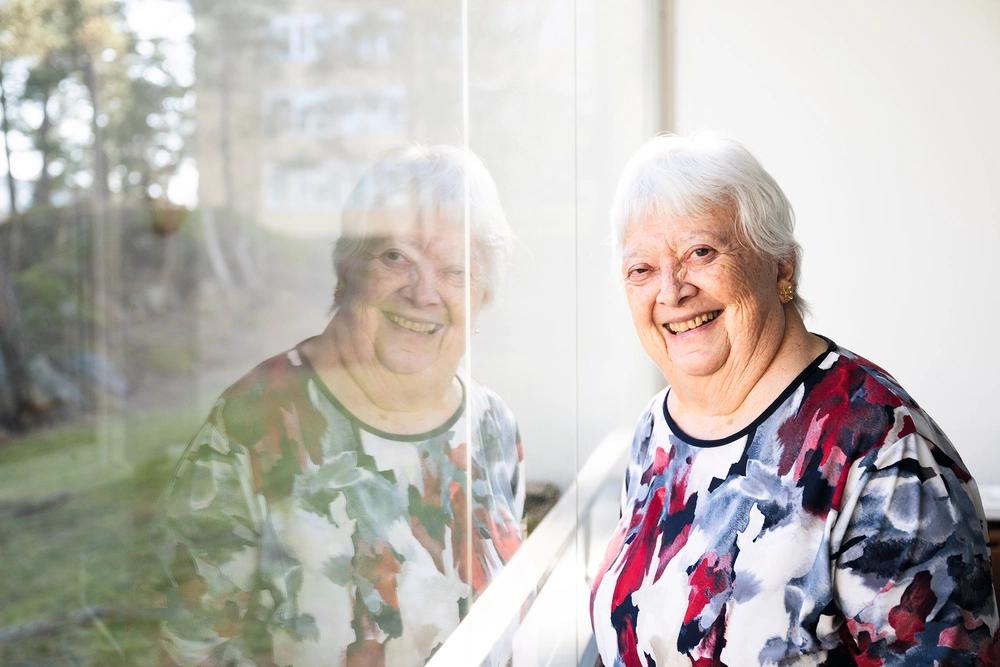 Leende äldre kvinna står vid ett fönster där hennes spegelbild syns i glaset.