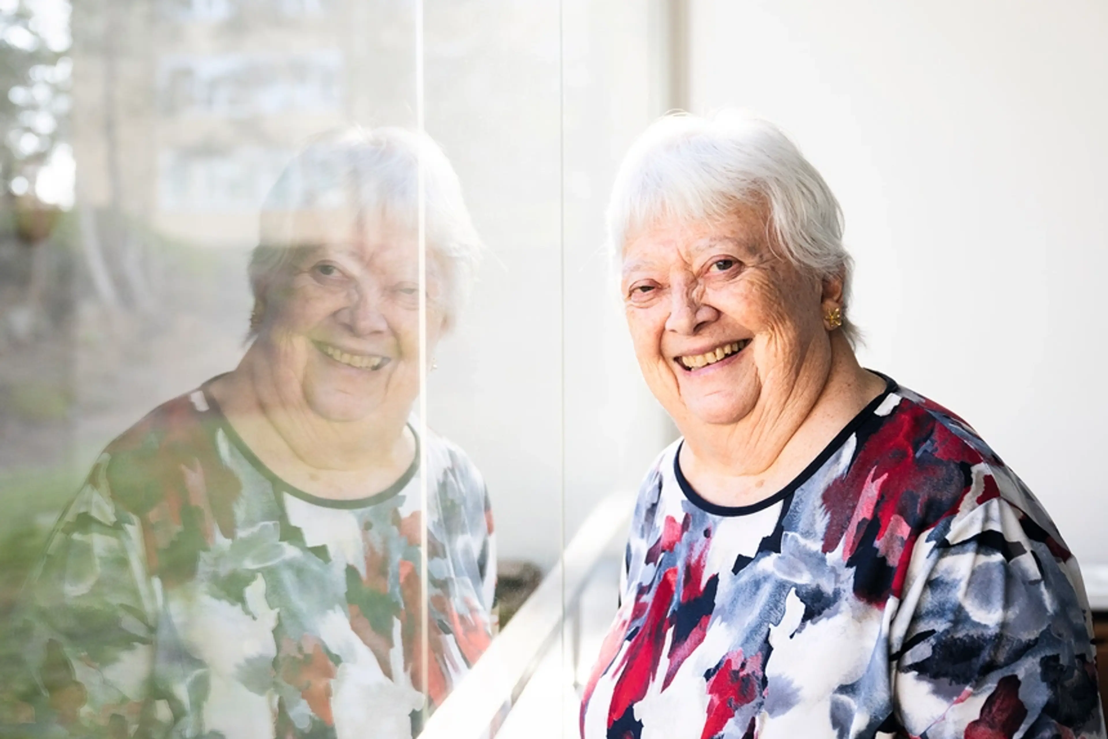 Leende äldre kvinna står vid ett fönster där hennes spegelbild syns i glaset.
