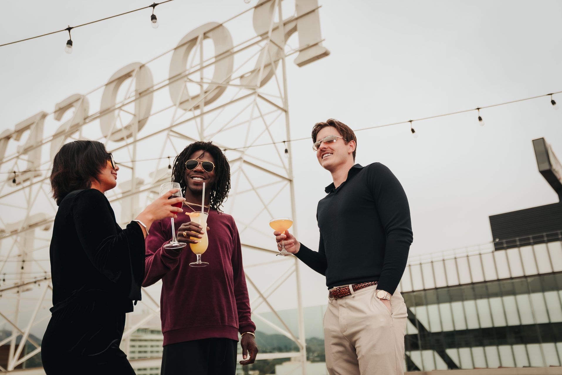 Three people are standing on a rooftop, toasting with drinks in front of a large sign.