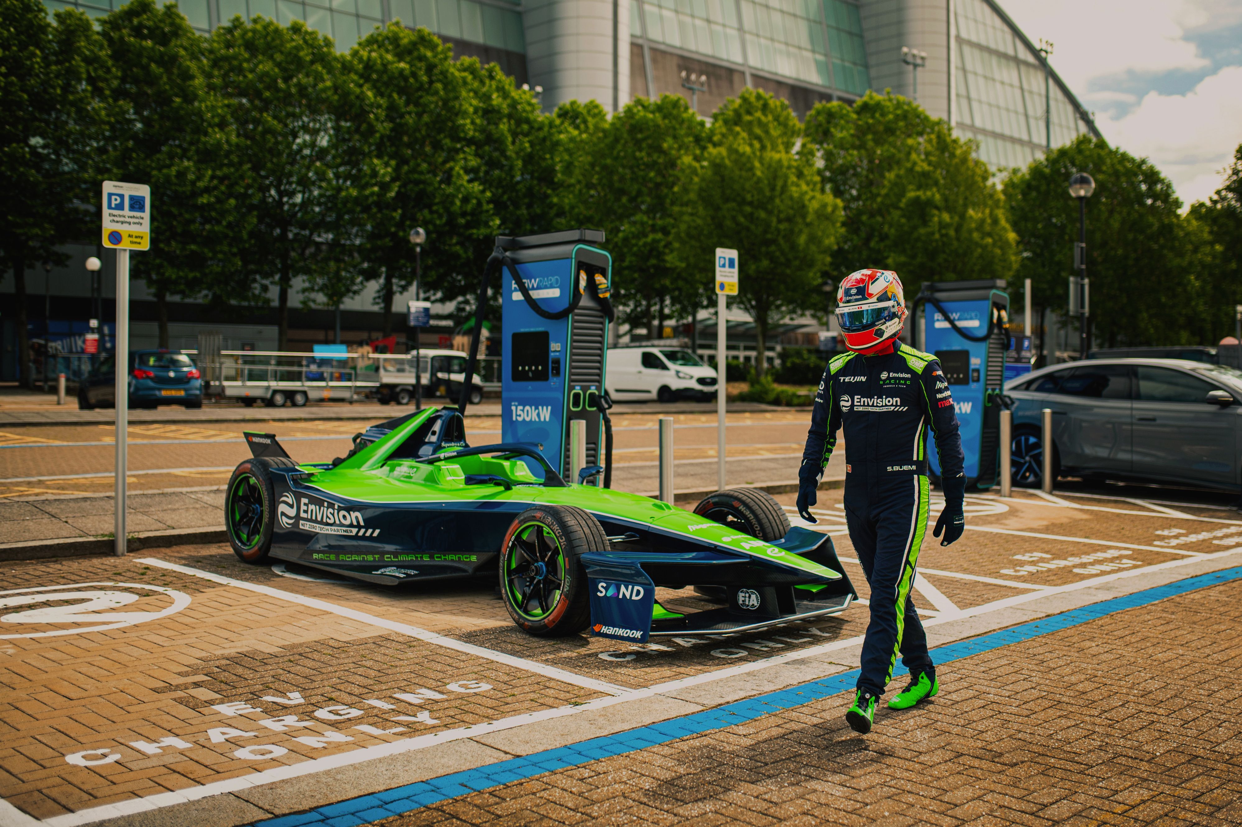 A racing driver in a black and green race kit walks away from a green and black electric race car plugged into a RAW charger