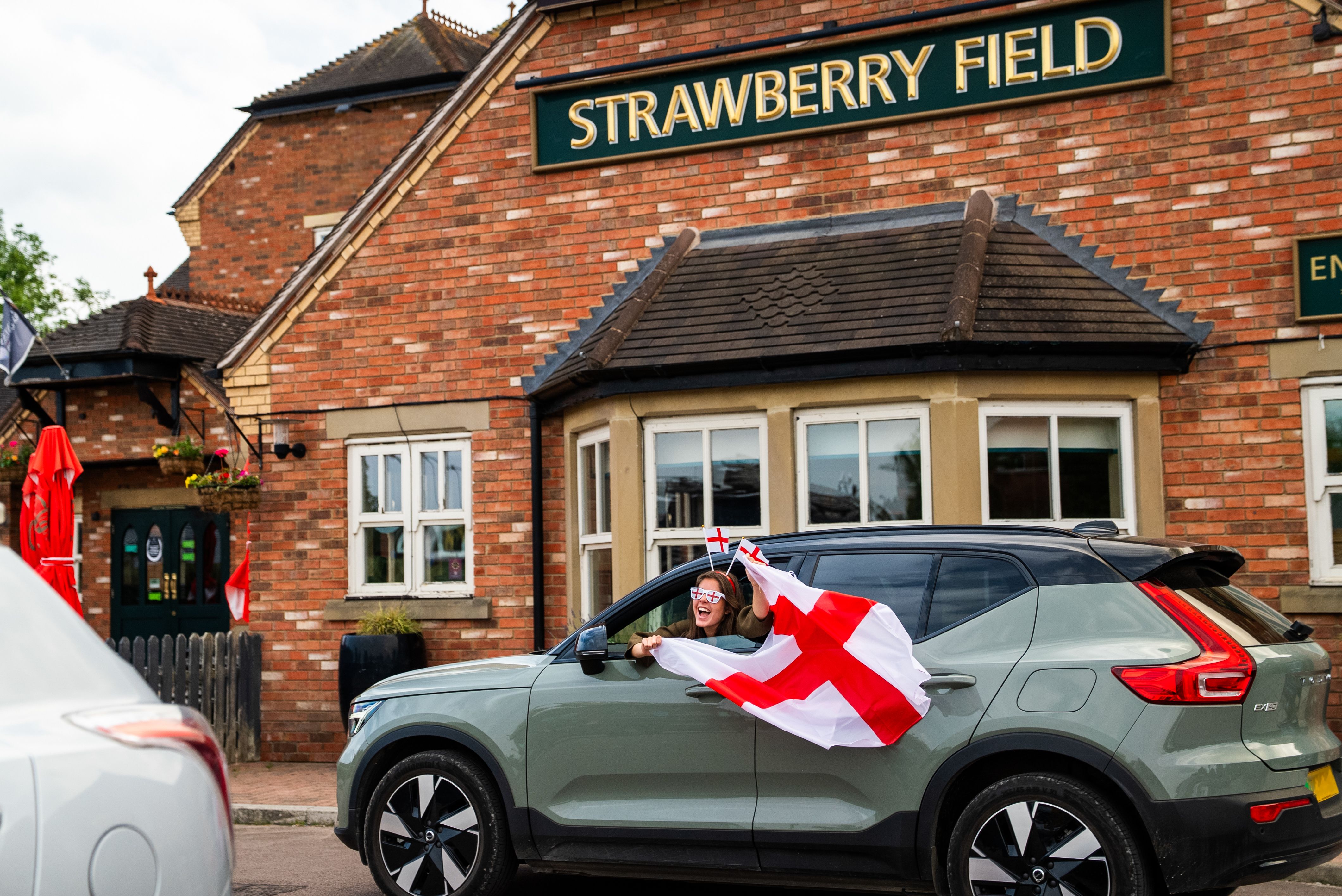 A woman wearing England flag sunglasses and headband waves an England flag out of a car window, outside the Greene King pub, Strawberry Field
