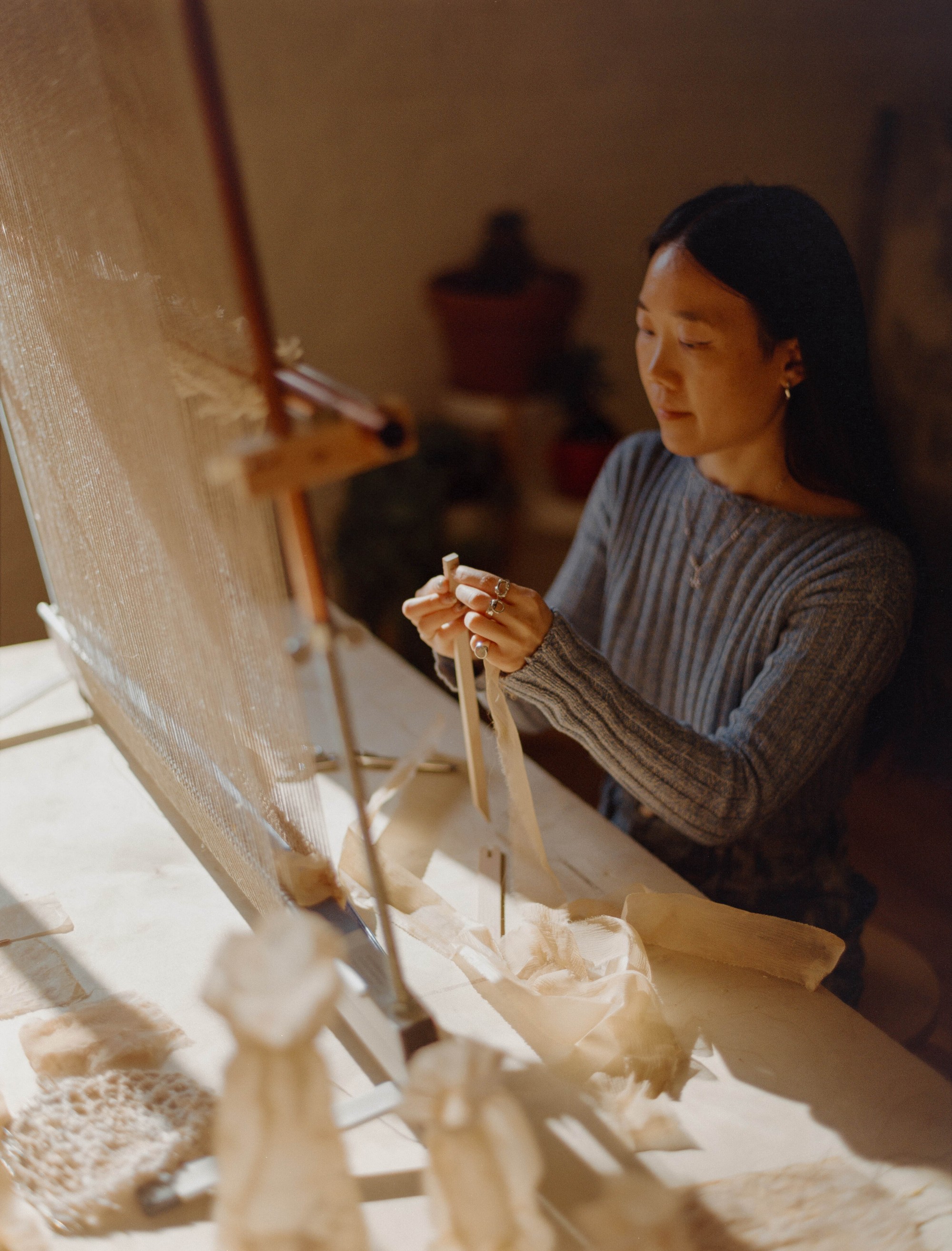Laura Kung threading fiber at her loom in her Portland studio, woven and dyed works around her, medium format film by Nathaniel Perales