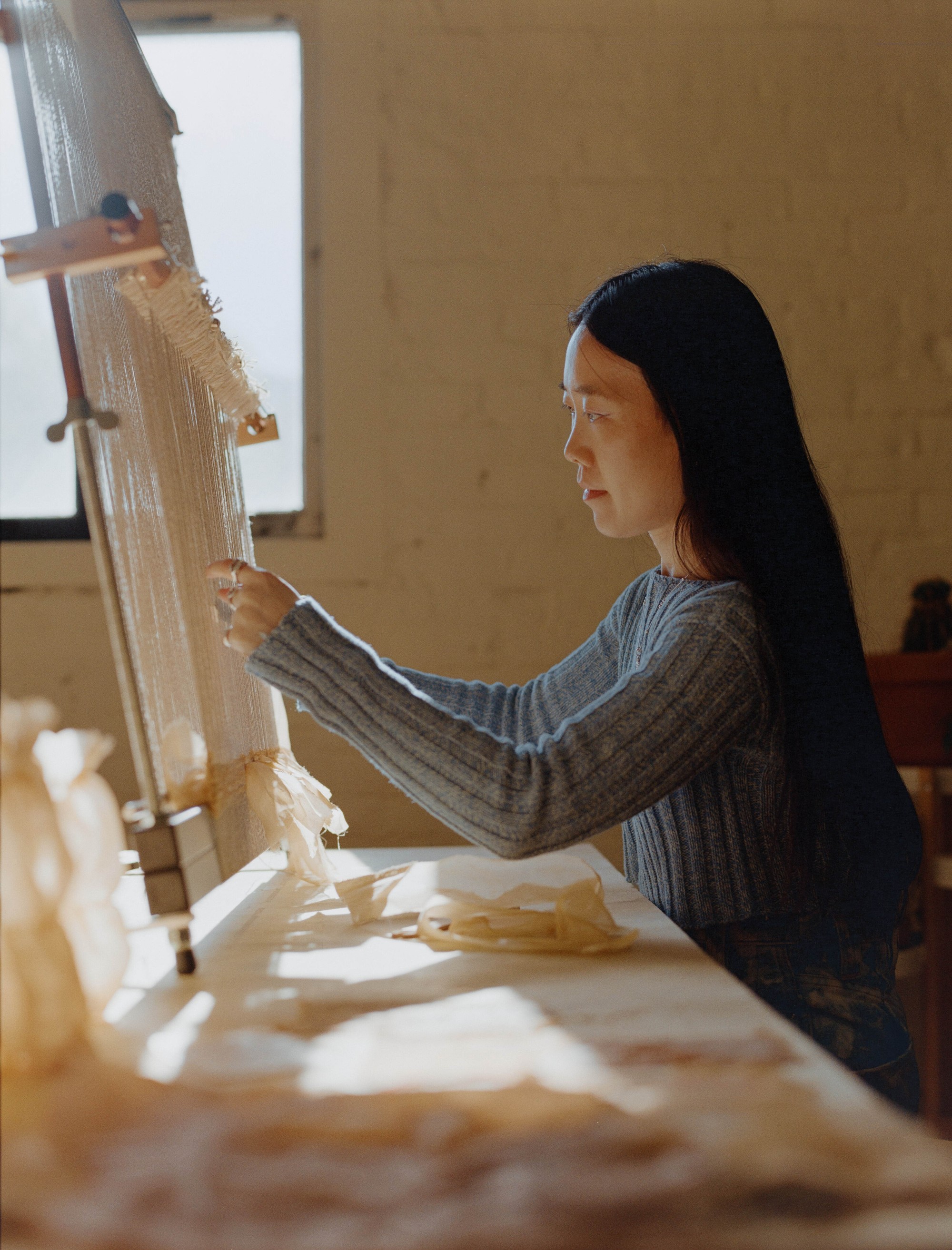 Laura Kung weaving at her loom in profile beside the studio window, Portland, shot on medium format film by Nathaniel Perales