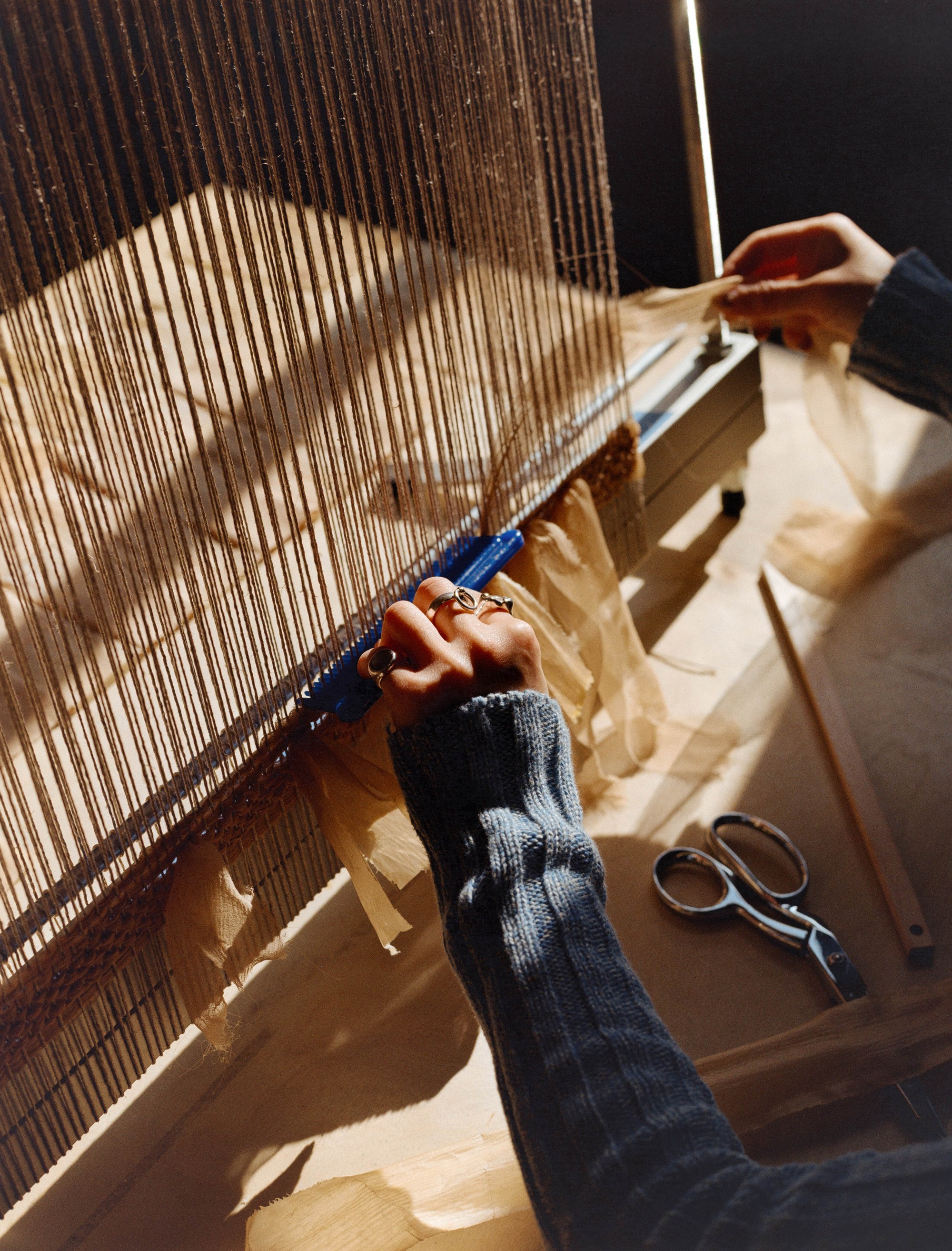 Laura Kung's hands working a blue weaving tool through warp threads beside scissors on the loom, Portland studio, medium format film by Nathaniel Perales