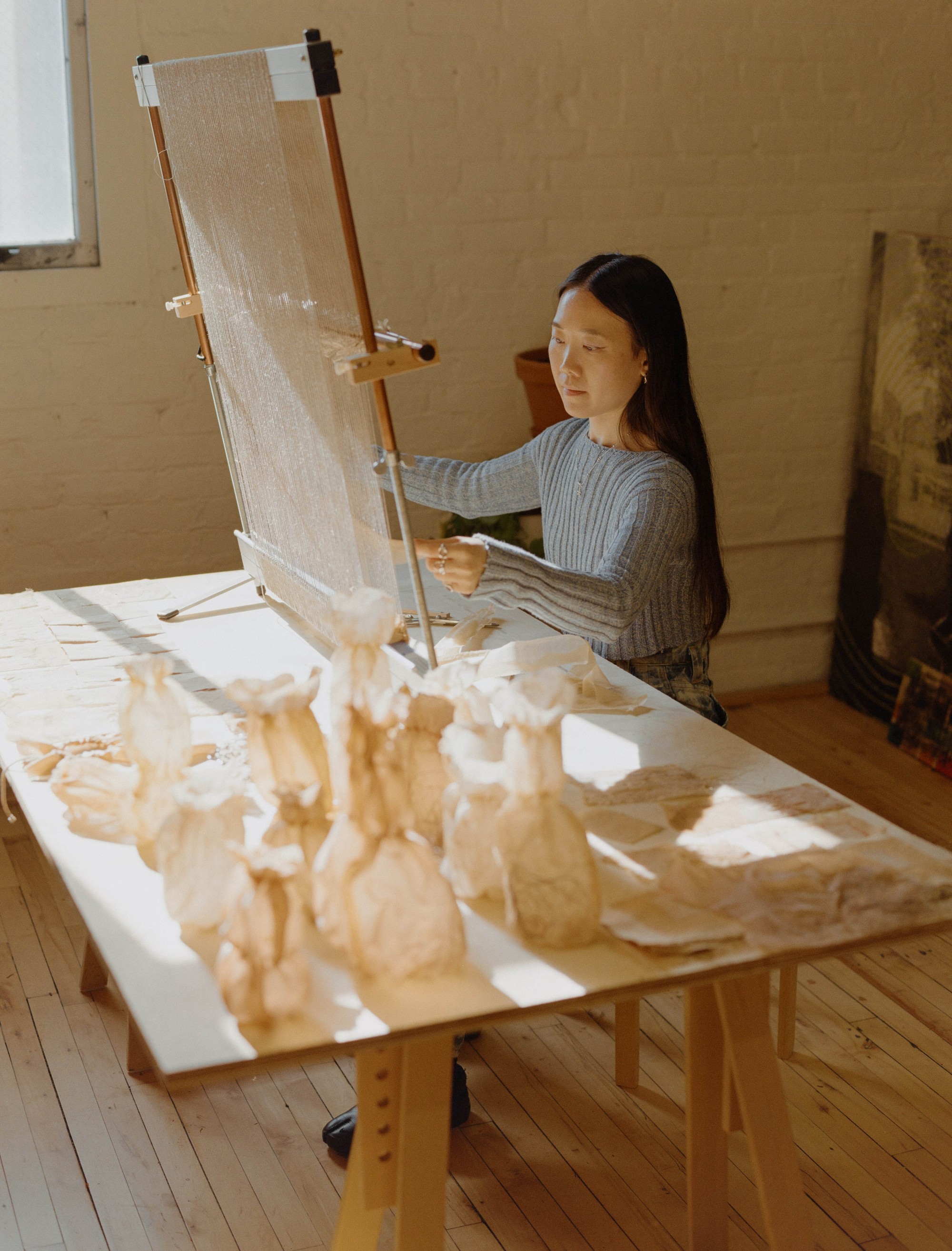 Laura Kung weaving at her tapestry loom with dyed fabric vessels on the table before her, Portland studio, medium format film by Nathaniel Perales