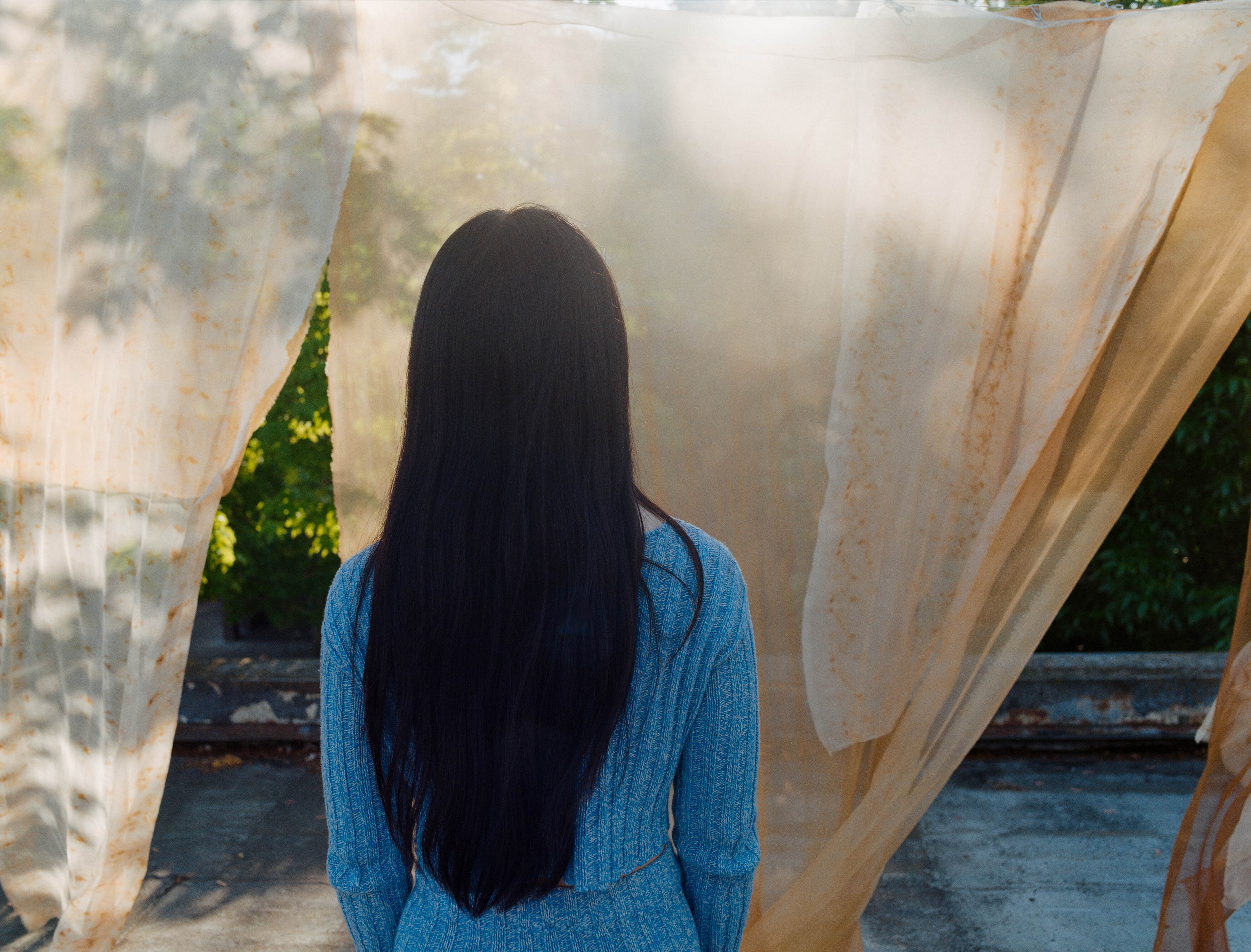Portland artist Laura Kung seen from behind between sunlit panels of naturally dyed fabric on a rooftop, shot on medium format film by Nathaniel Perales