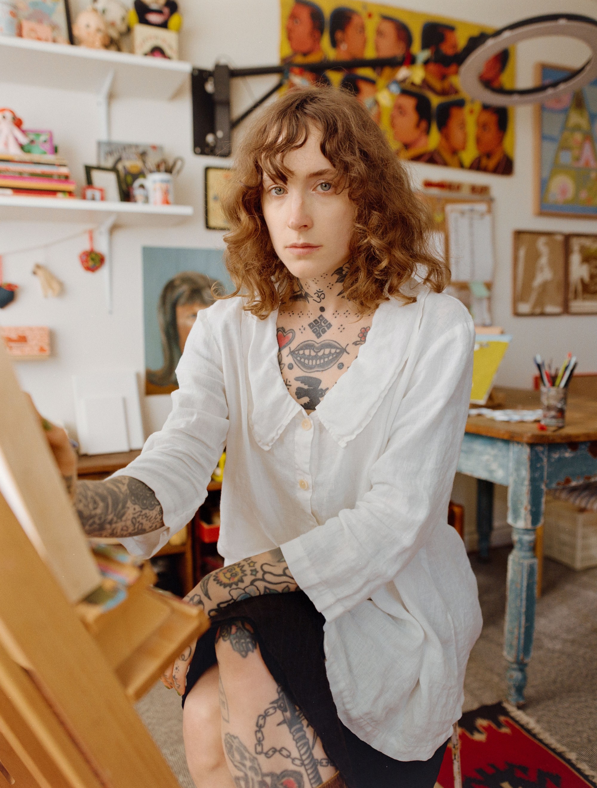 Addeline Griswold painting at her easel in her Los Angeles studio, art and collectibles visible throughout the room, medium format film portrait by Nathaniel Perales