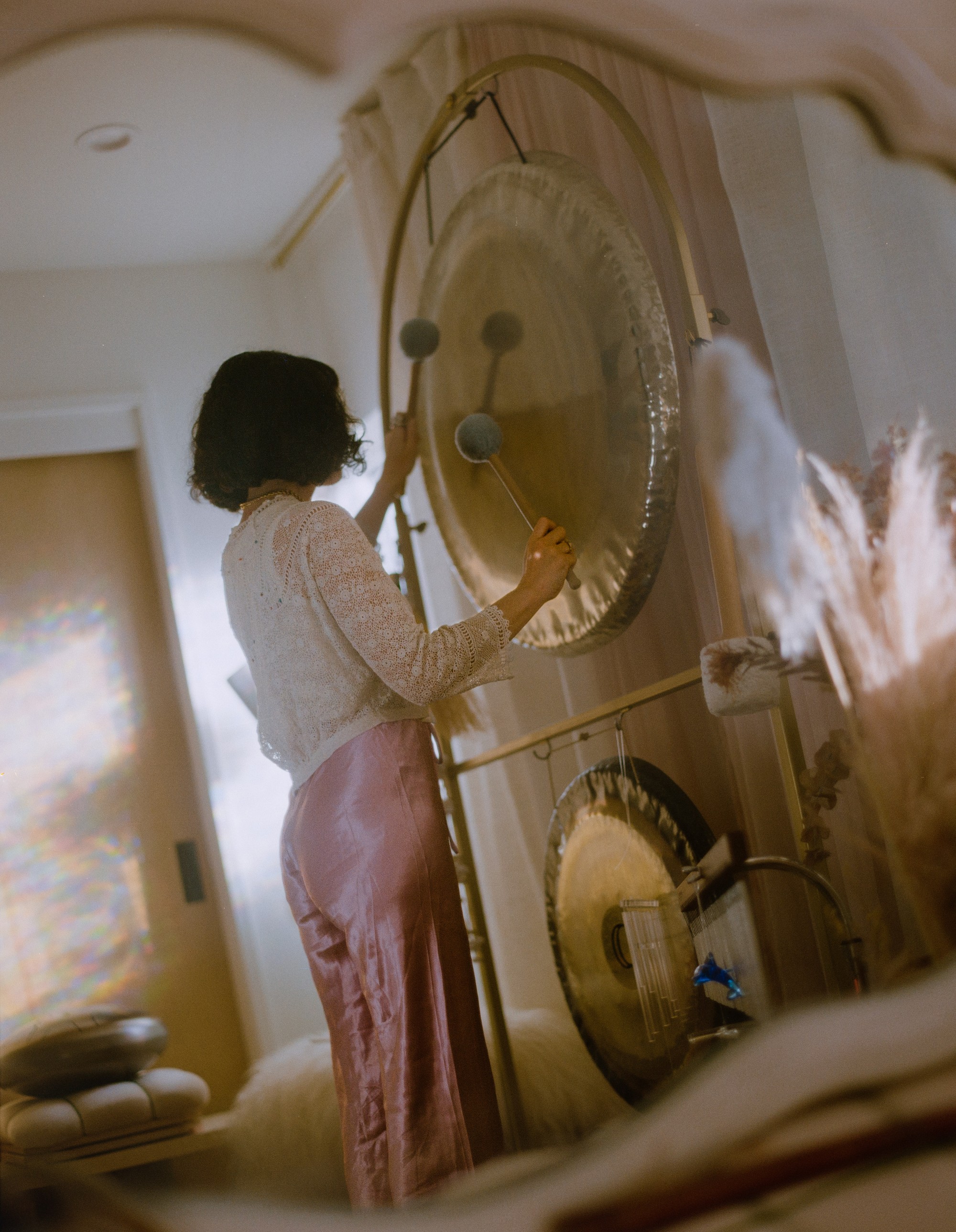 Allison Bagg playing her large gong from behind in her Los Angeles home, rainbow prism light visible, shot on medium format film