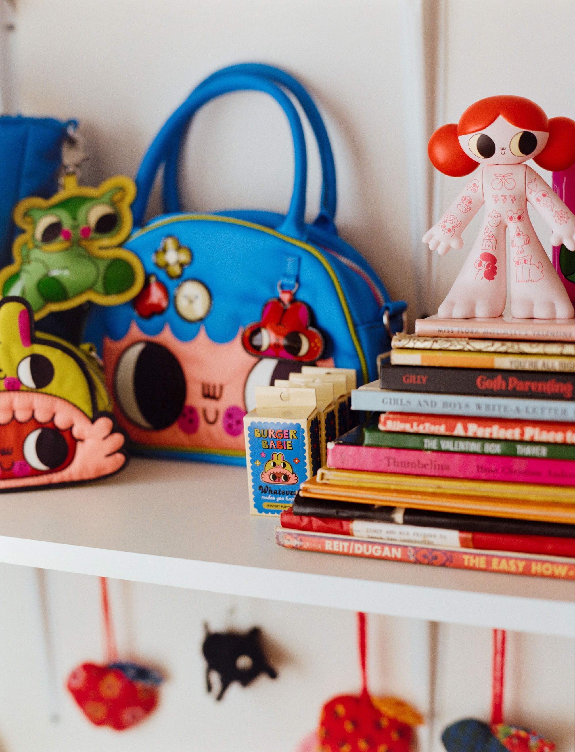 Shelf detail in Addeline Griswold's studio showing Burger Babie merchandise, designer toys, and stacked books, photographed on film by Nathaniel Perales