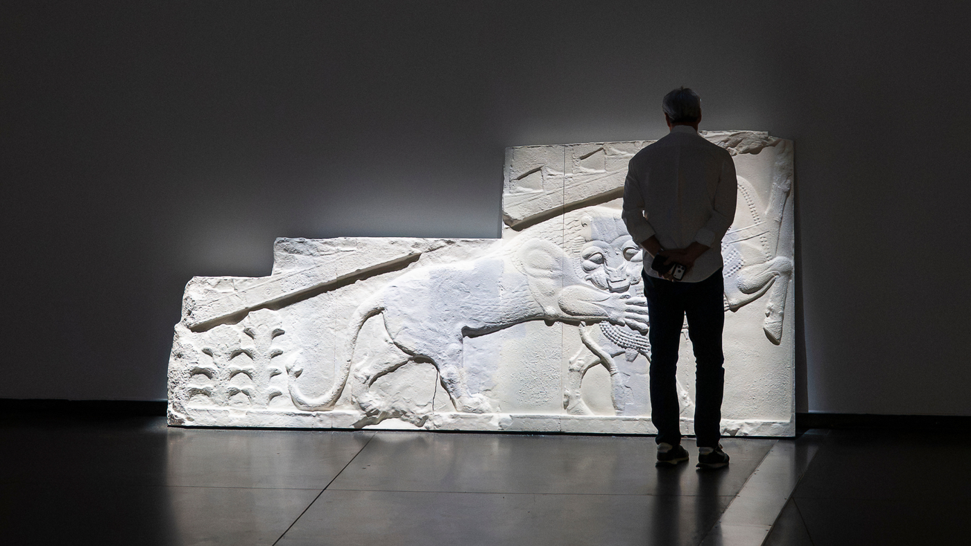 A person in a white shirt stands with their back to the camera, hands clasped behind them, looking at a large white relief sculpture of a big cat leaning against a dark gallery wall, lit from above.