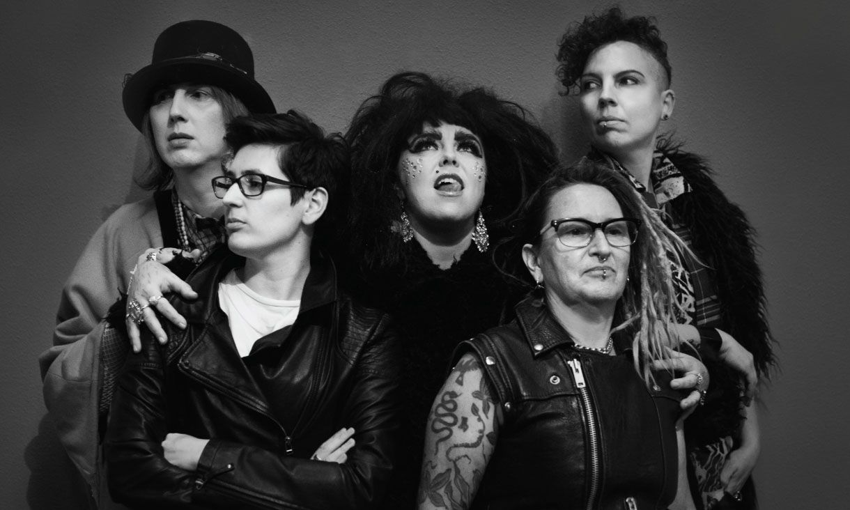 Black-and-white photo of five people posed close together against a plain wall. At the centre, one person with big dark hair and gems on their face looks upward, mouth open.