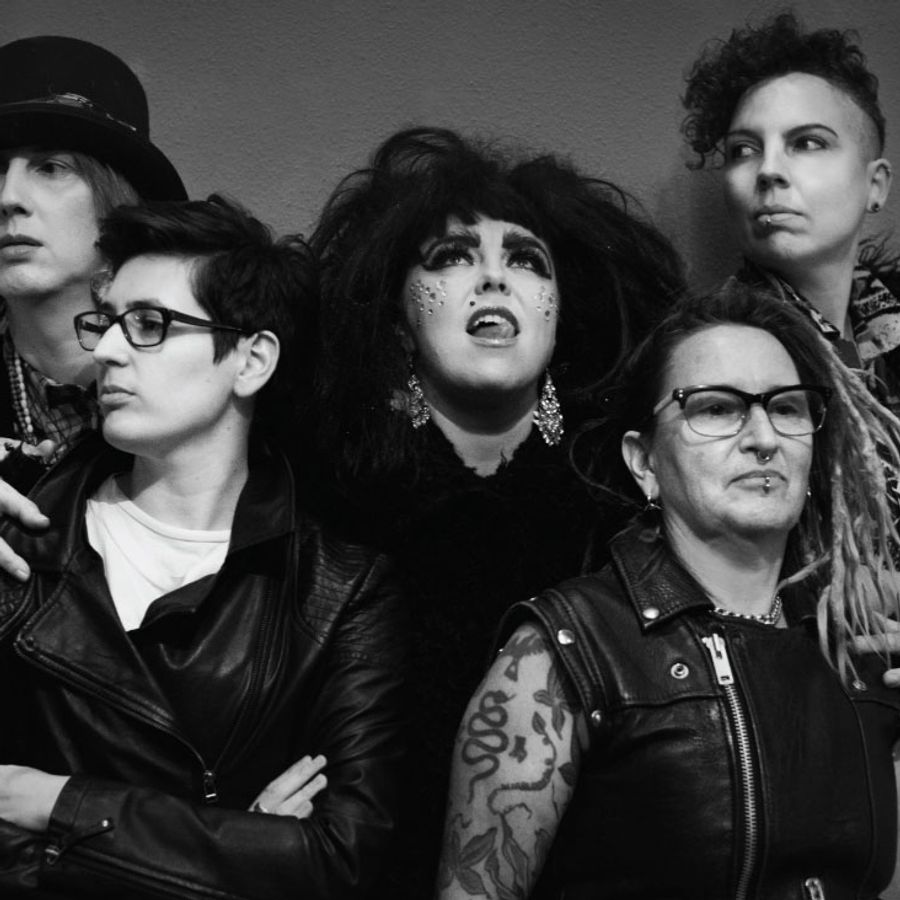 Black-and-white photo of five people posed close together against a plain wall. At the centre, one person with big dark hair and gems on their face looks upward, mouth open.