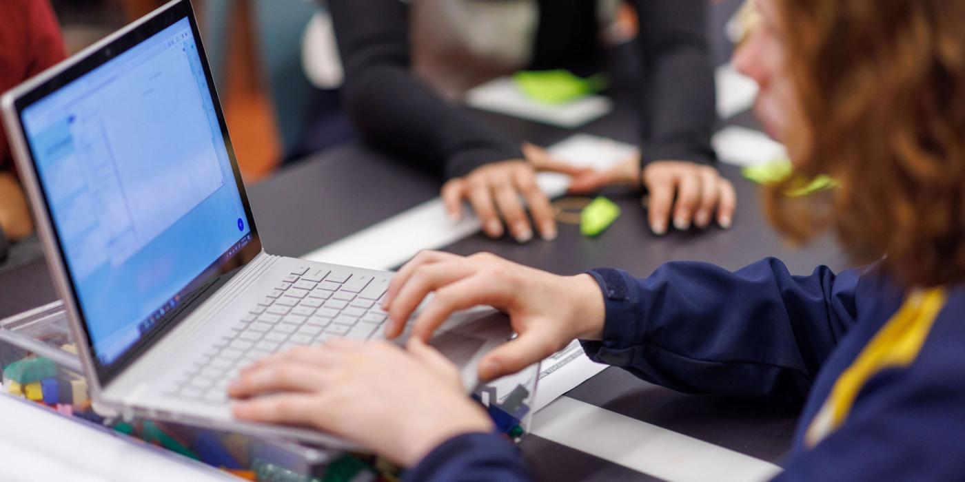 A young person types on a laptop at a workshop table, with other participants’ hands and coloured building blocks in the background.