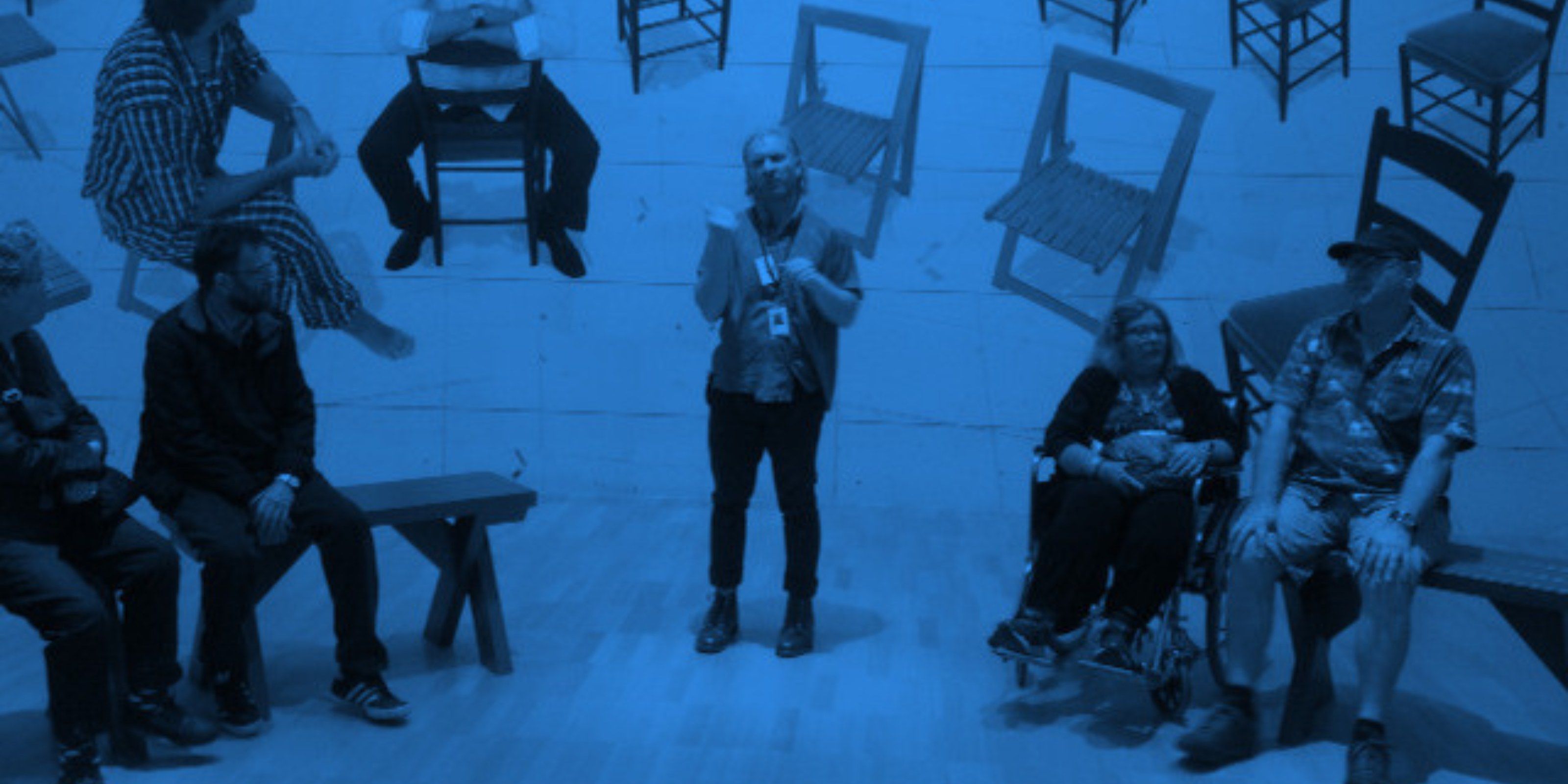 Blue-toned photo of a guide wearing a lanyard, standing in a gallery and talking with a group seated on benches, one in a wheelchair. Wooden chairs are mounted on the wall behind them.