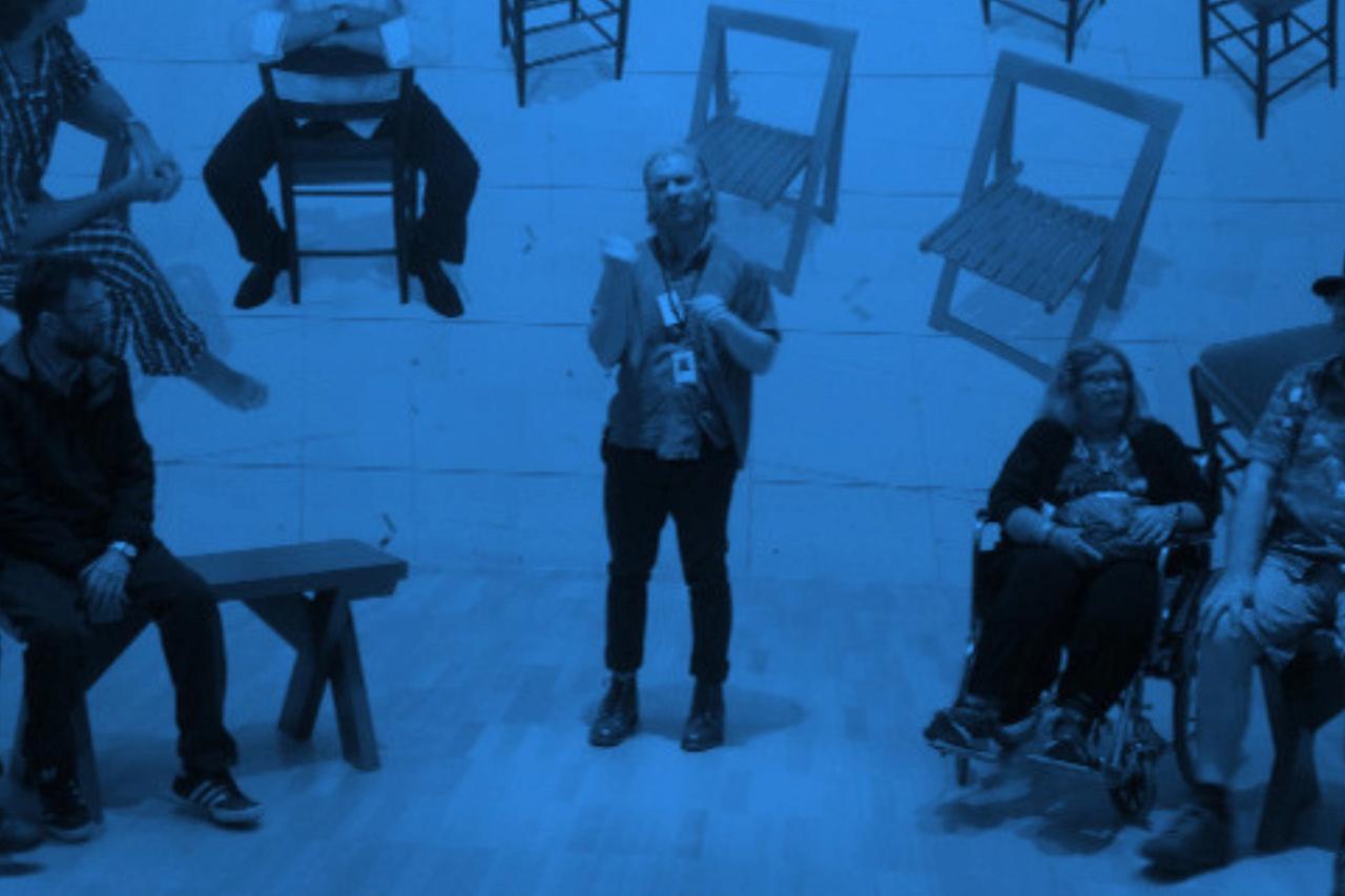 Blue-toned photo of a guide wearing a lanyard, standing in a gallery and talking with a group seated on benches, one in a wheelchair. Wooden chairs are mounted on the wall behind them.